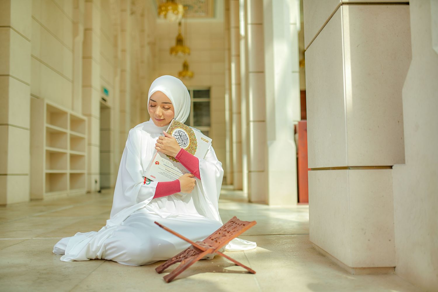 Muslim woman in white hijab and prayer robe reading Quran on decorative wooden stand indoors.