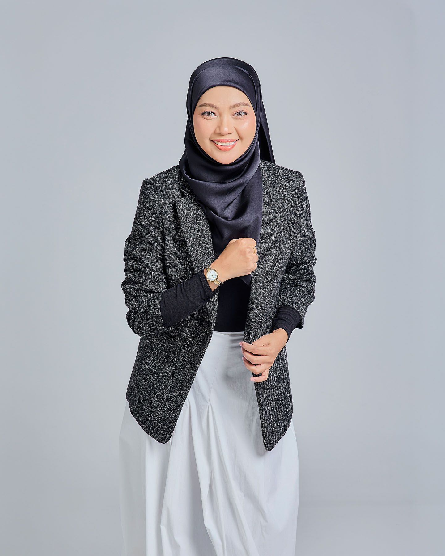 Muslim woman in gray herringbone blazer, white skirt, and navy hijab smiling while adjusting her sleeve.