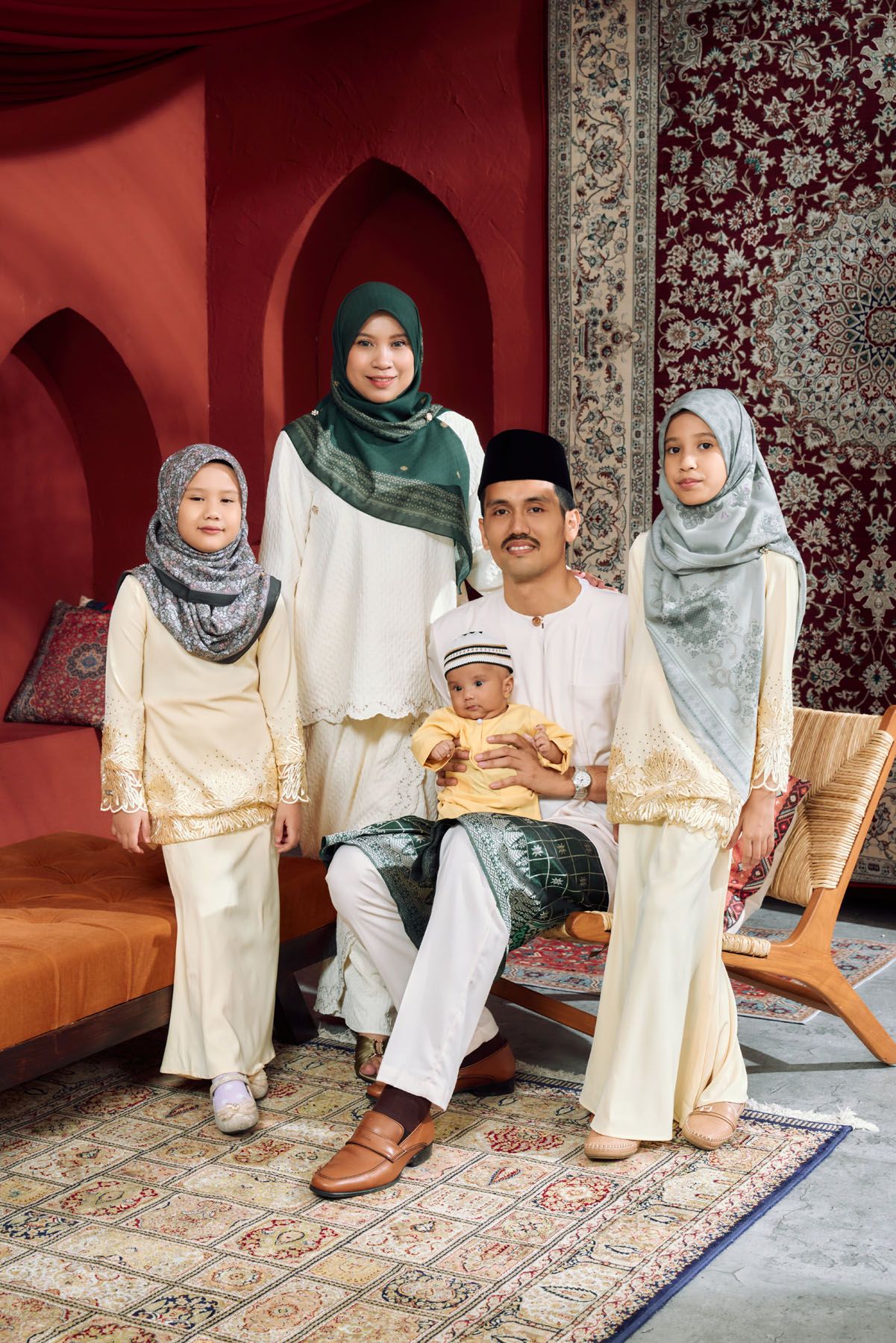 Muslim family in cream traditional Malay attire with father holding baby, two daughters in hijabs for Eid celebration.