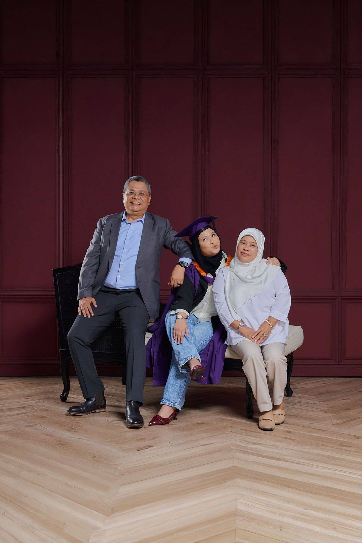 Graduate in purple gown sitting between parents against dark red paneled wall for portrait.