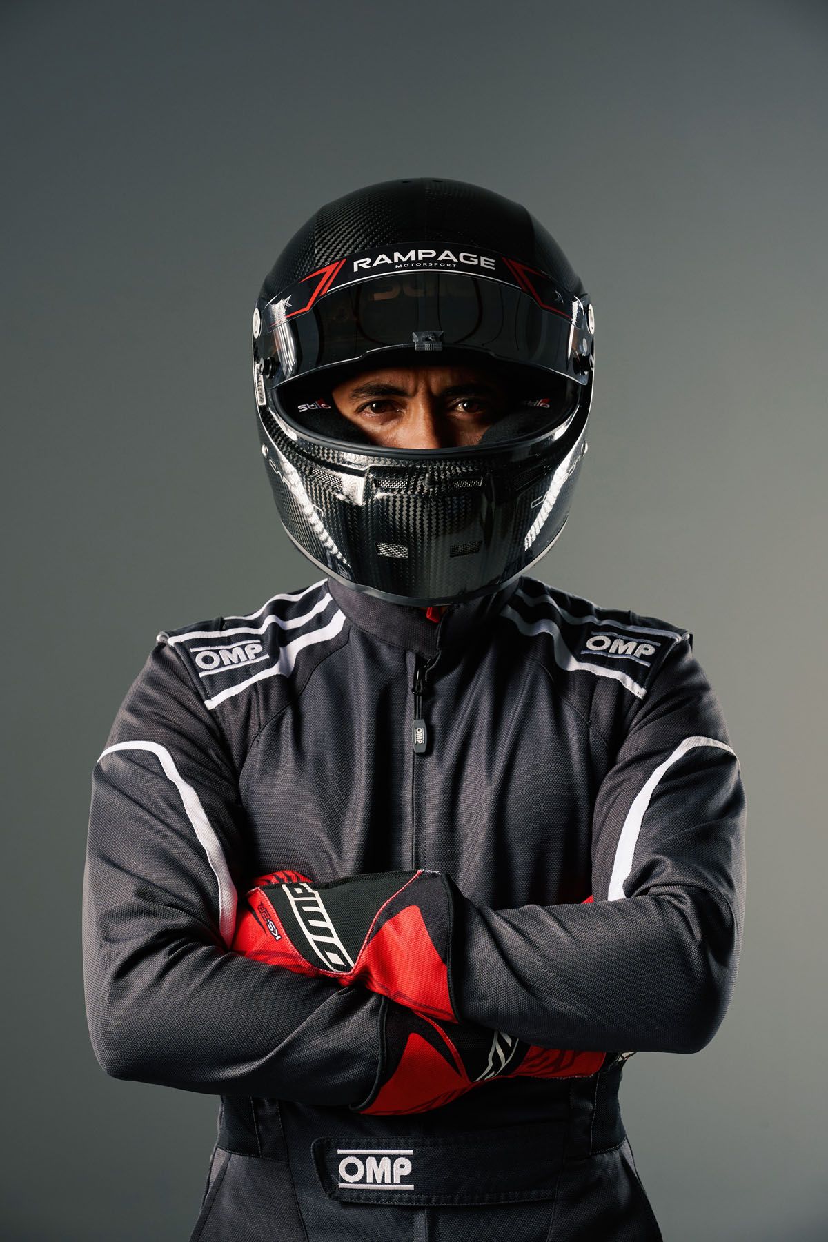 Racing driver in black OMP suit and Rampage helmet stands with arms crossed in a studio portrait.