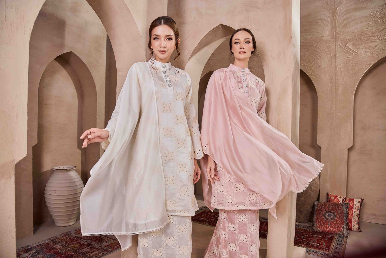 Two women modeling elegant South Asian embroidered outfits in cream and pink tones in an arched setting.