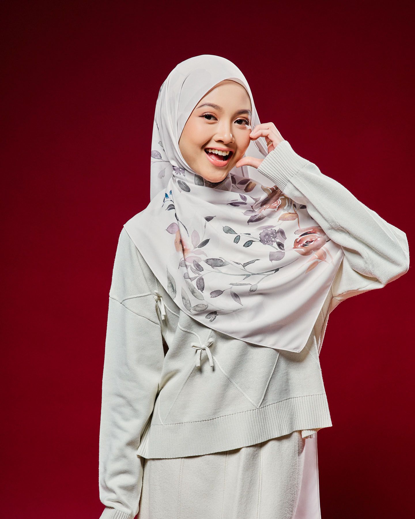 Smiling woman in white floral hijab and cream outfit poses against red background.