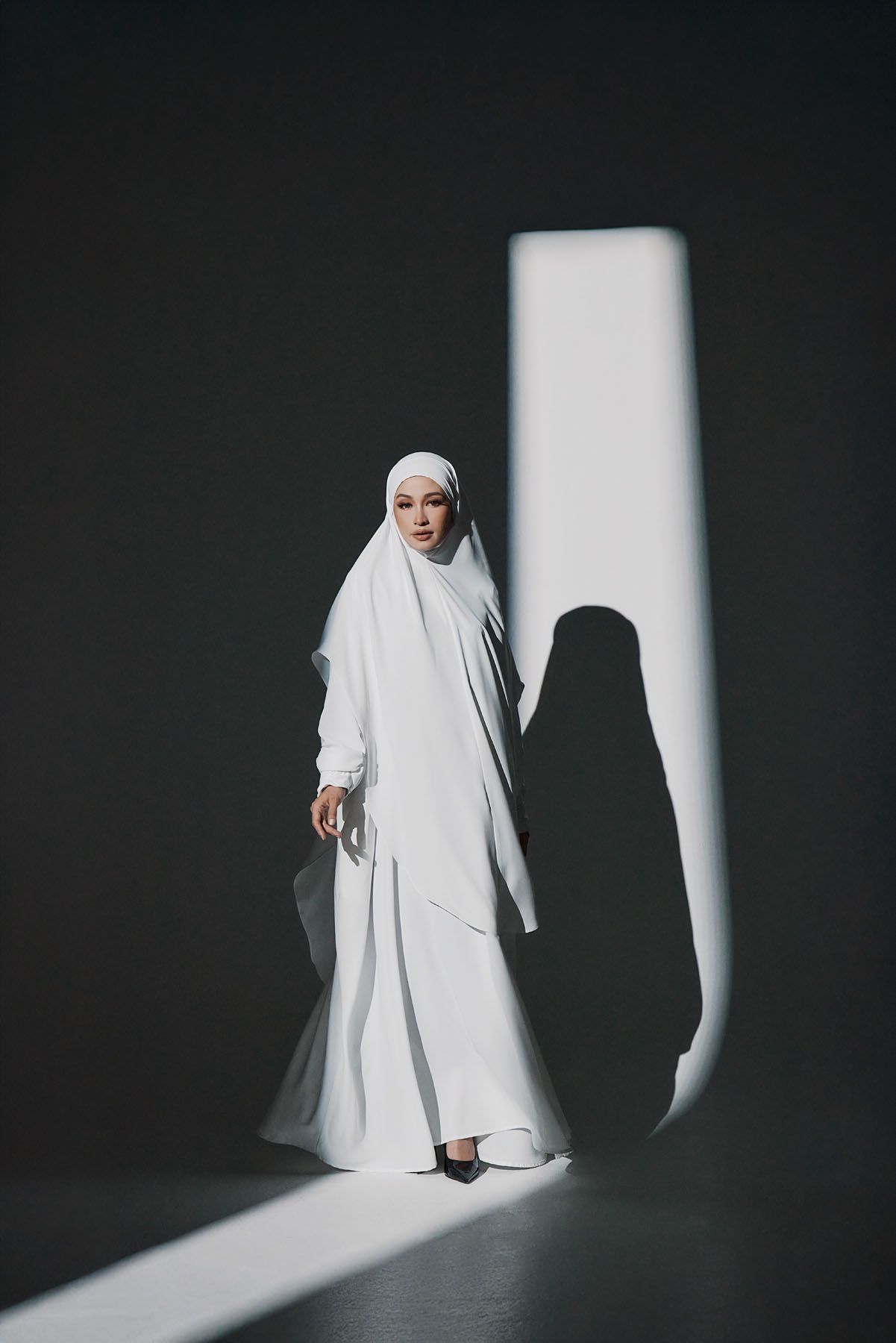 Muslim woman in flowing white hijab and abaya standing in dramatic light against dark background.
