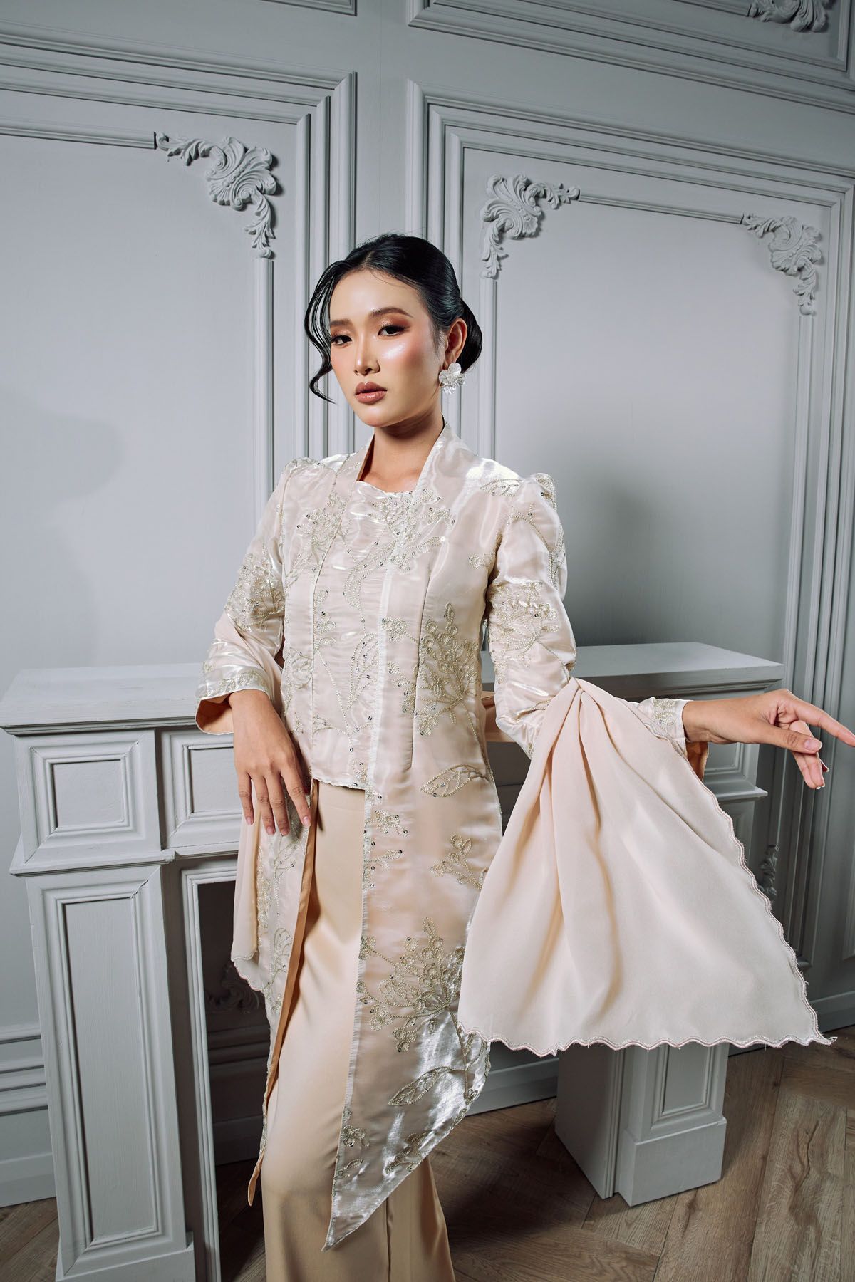 Woman in cream embroidered kebaya with gold detailing poses gracefully in a white classical interior setting.