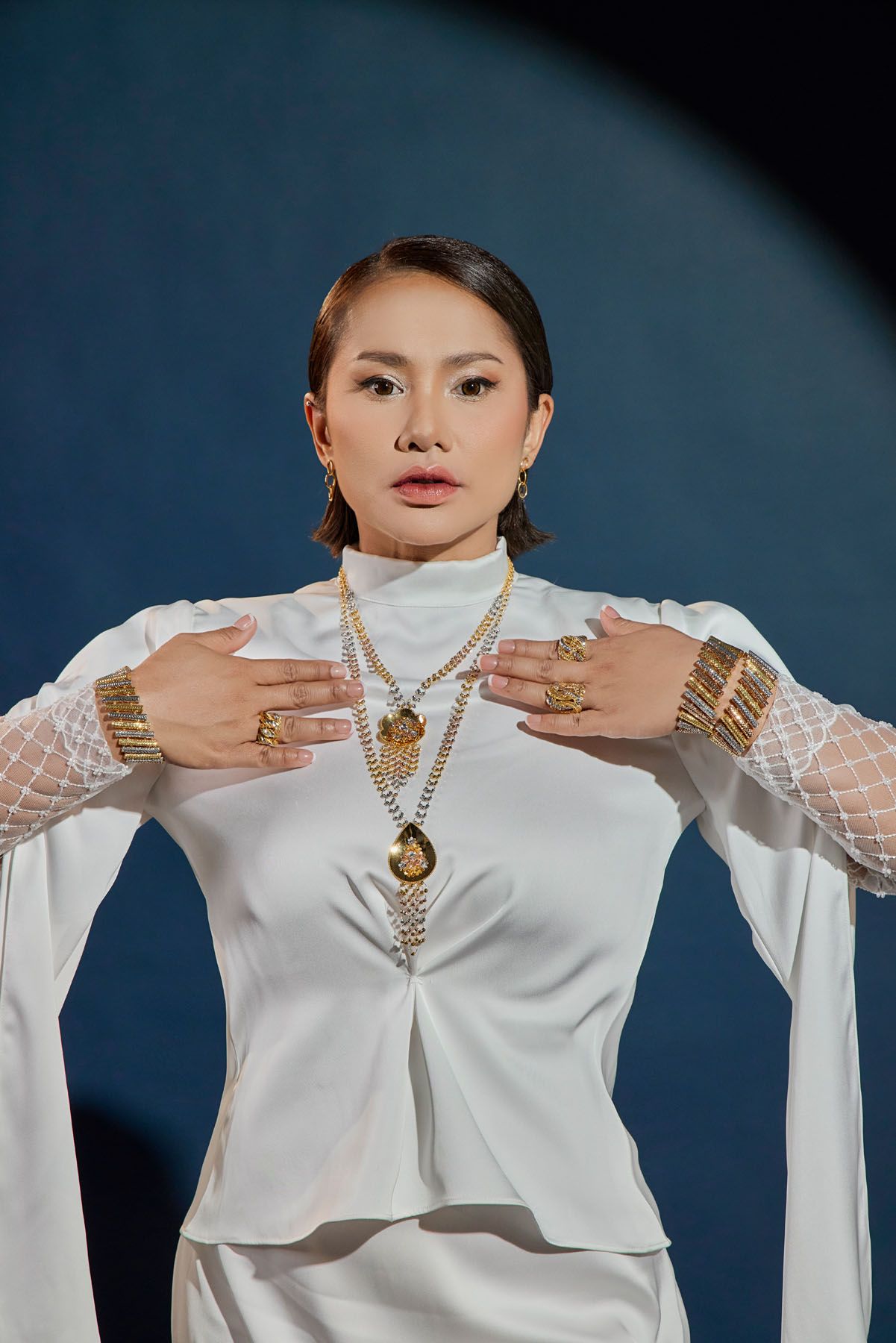 Woman in white high-neck outfit displaying gold jewelry necklaces rings and bracelets on blue background.