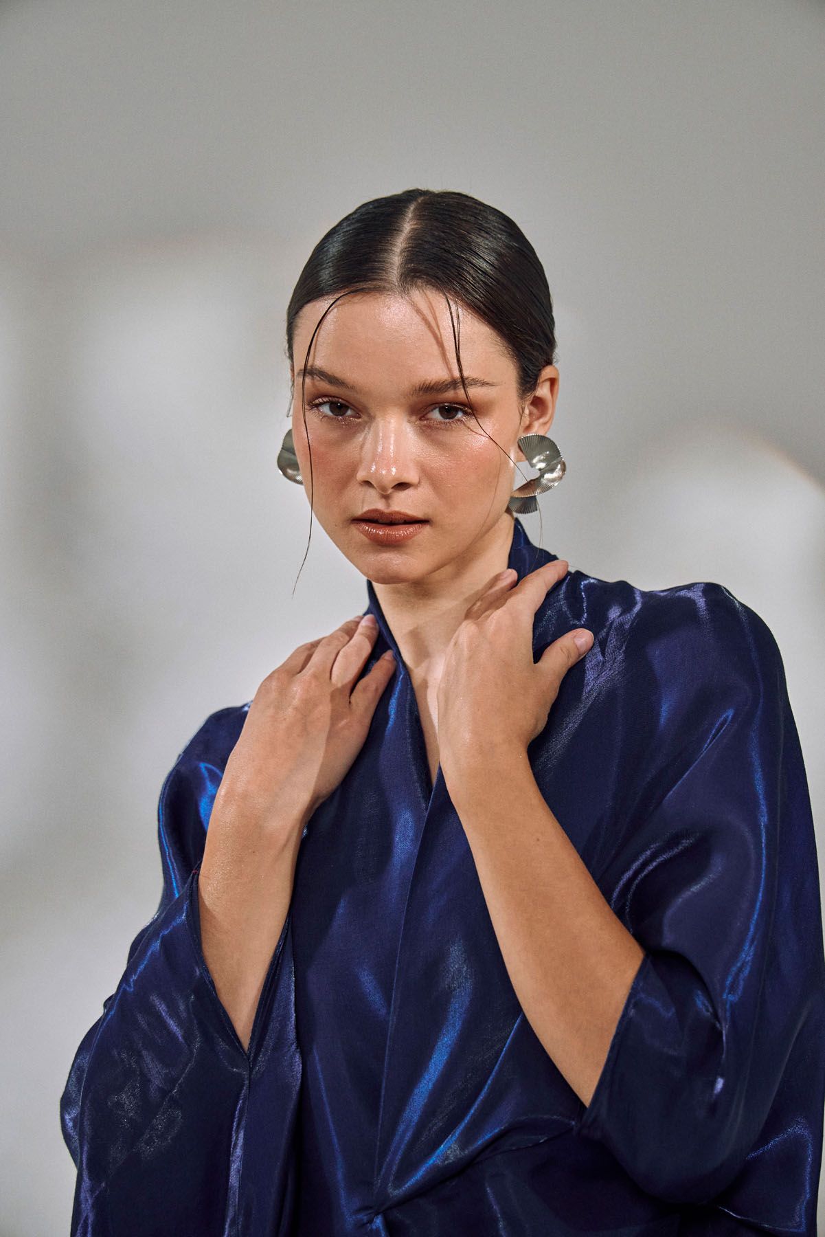 Woman in a deep blue satin kimono-style top adjusts collar, wearing silver earrings on gray background.