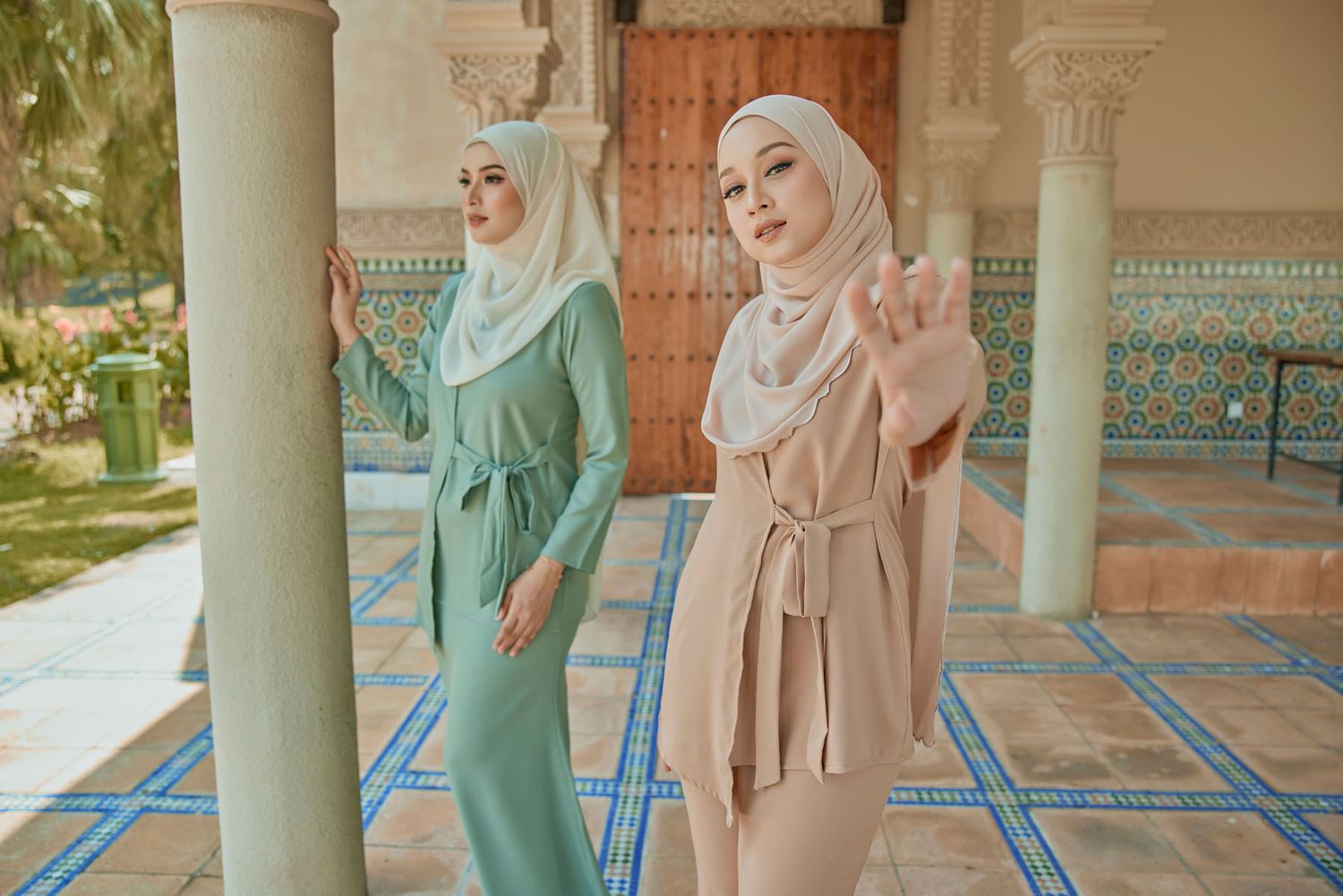 Two women in green and beige hijab outfits pose near ornate columns in an outdoor setting.