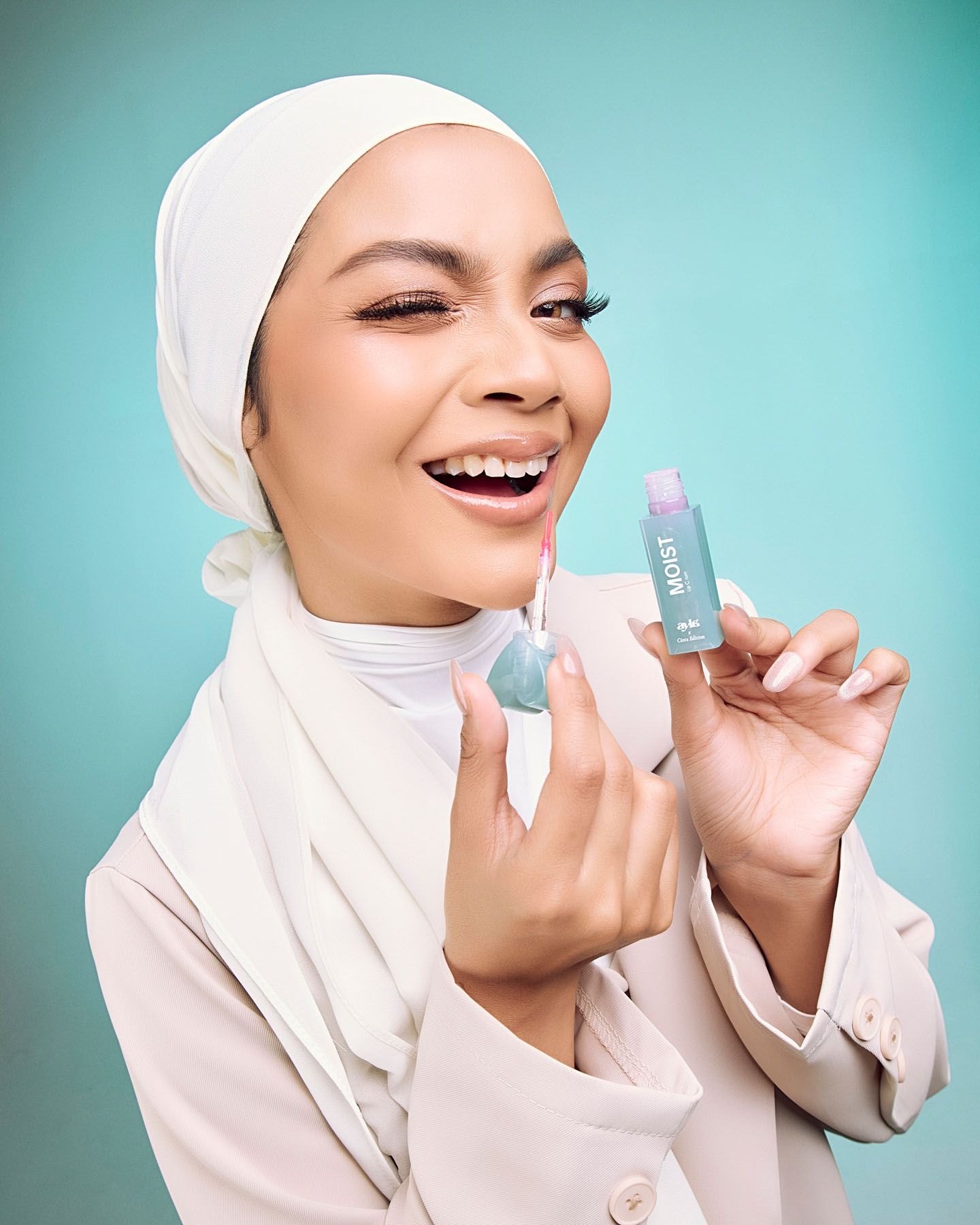 Joyful hijab woman holding light blue lip gloss product on turquoise background.