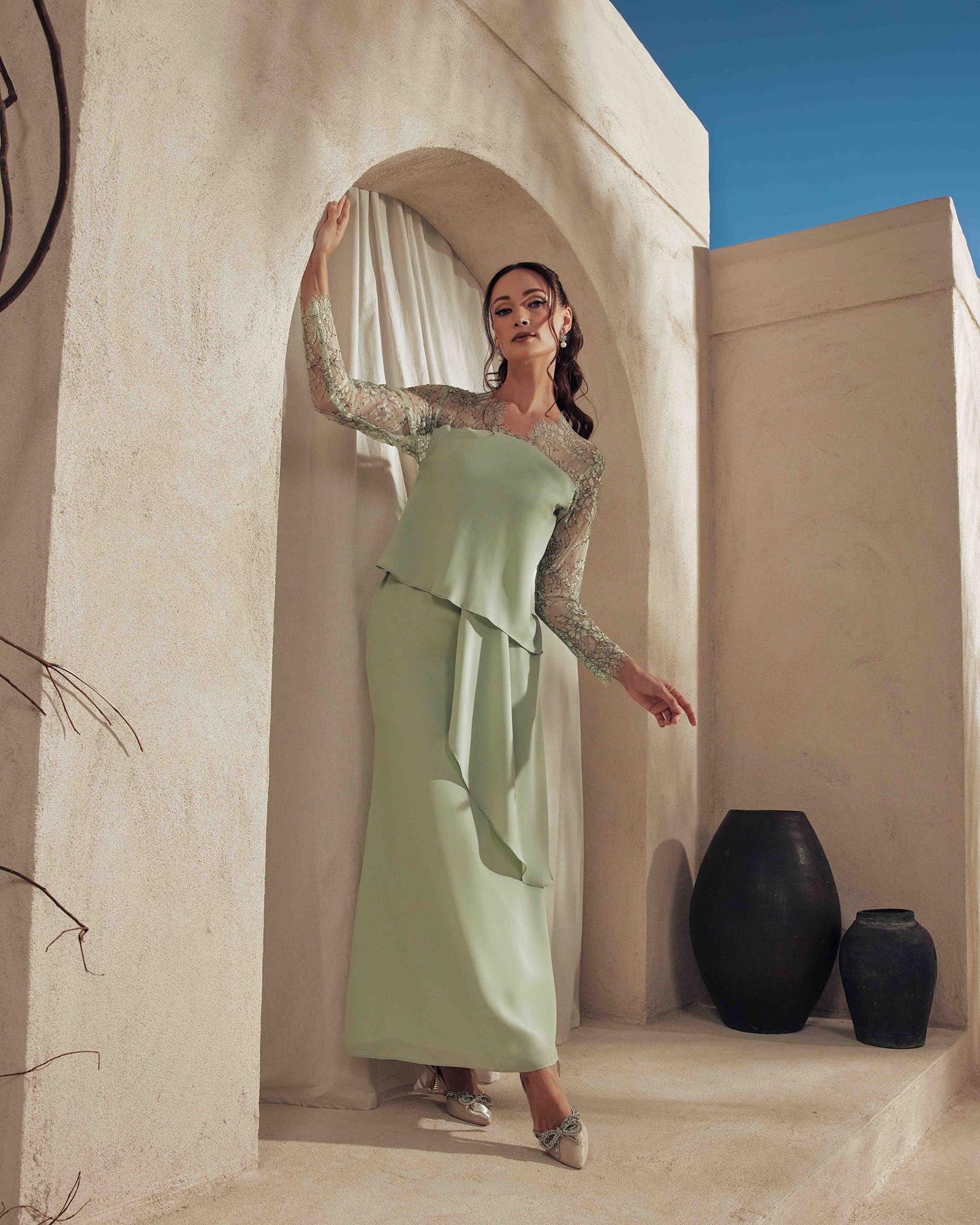 Woman in sage green modest dress with lace sleeves standing in Mediterranean style archway.