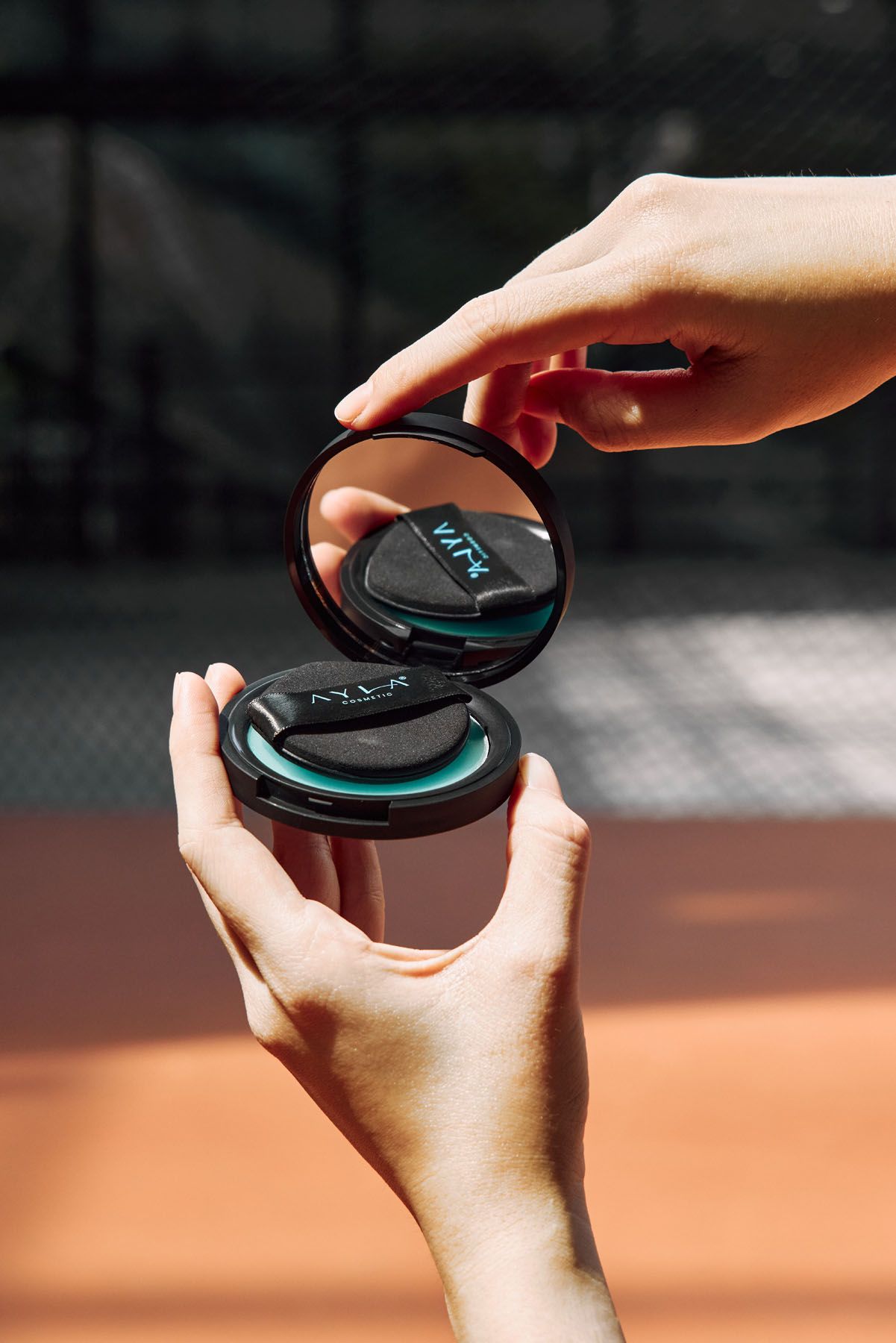 Hands holding open AYLA black compact makeup case with mirror on a sunlit clay court background.