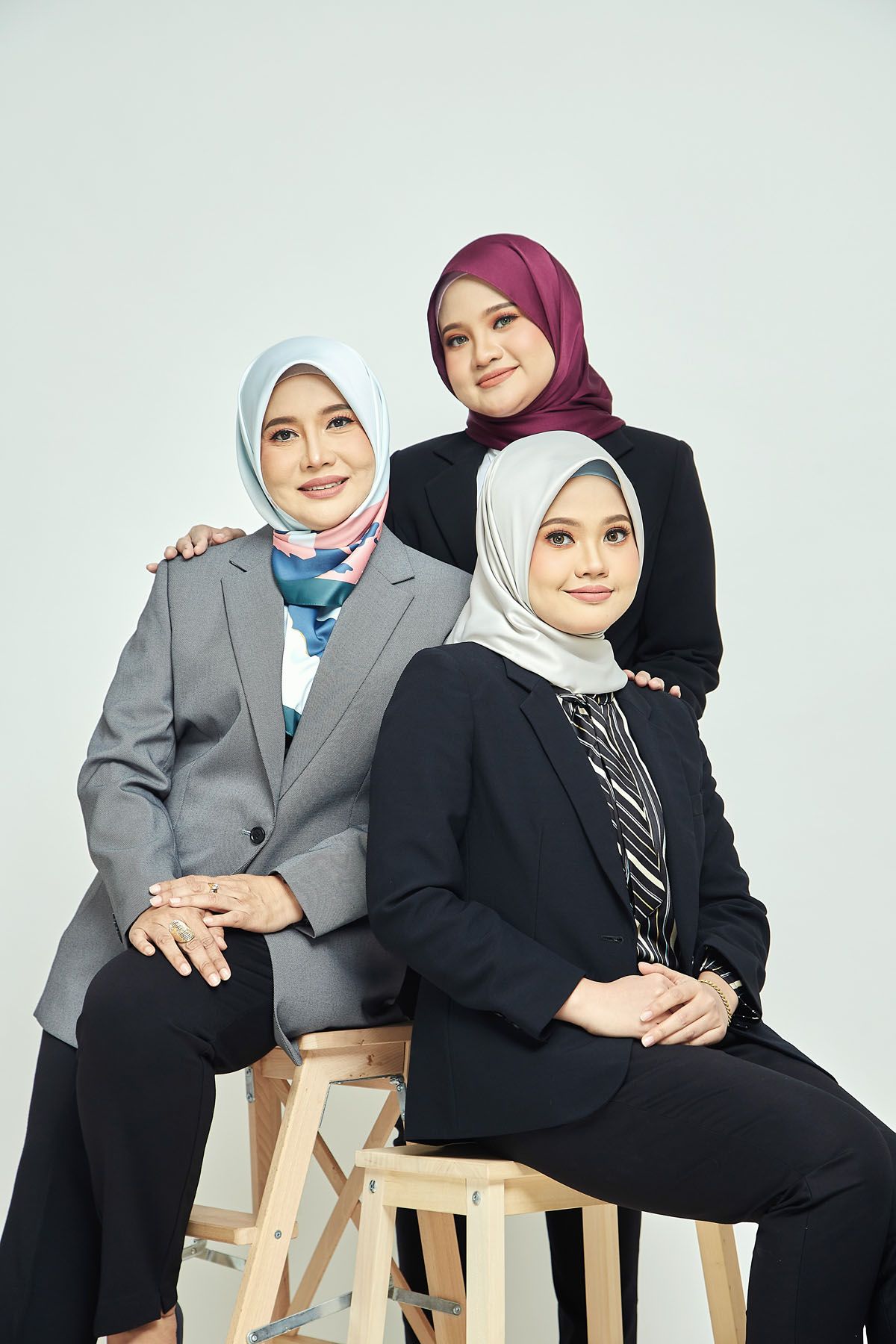 Three Muslim women in hijabs and business suits posing together in a professional studio photo.