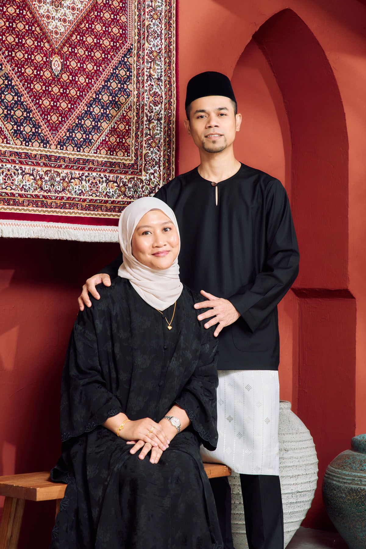Muslim mother in hijab seated with son in baju melayu posing before decorative carpet backdrop.