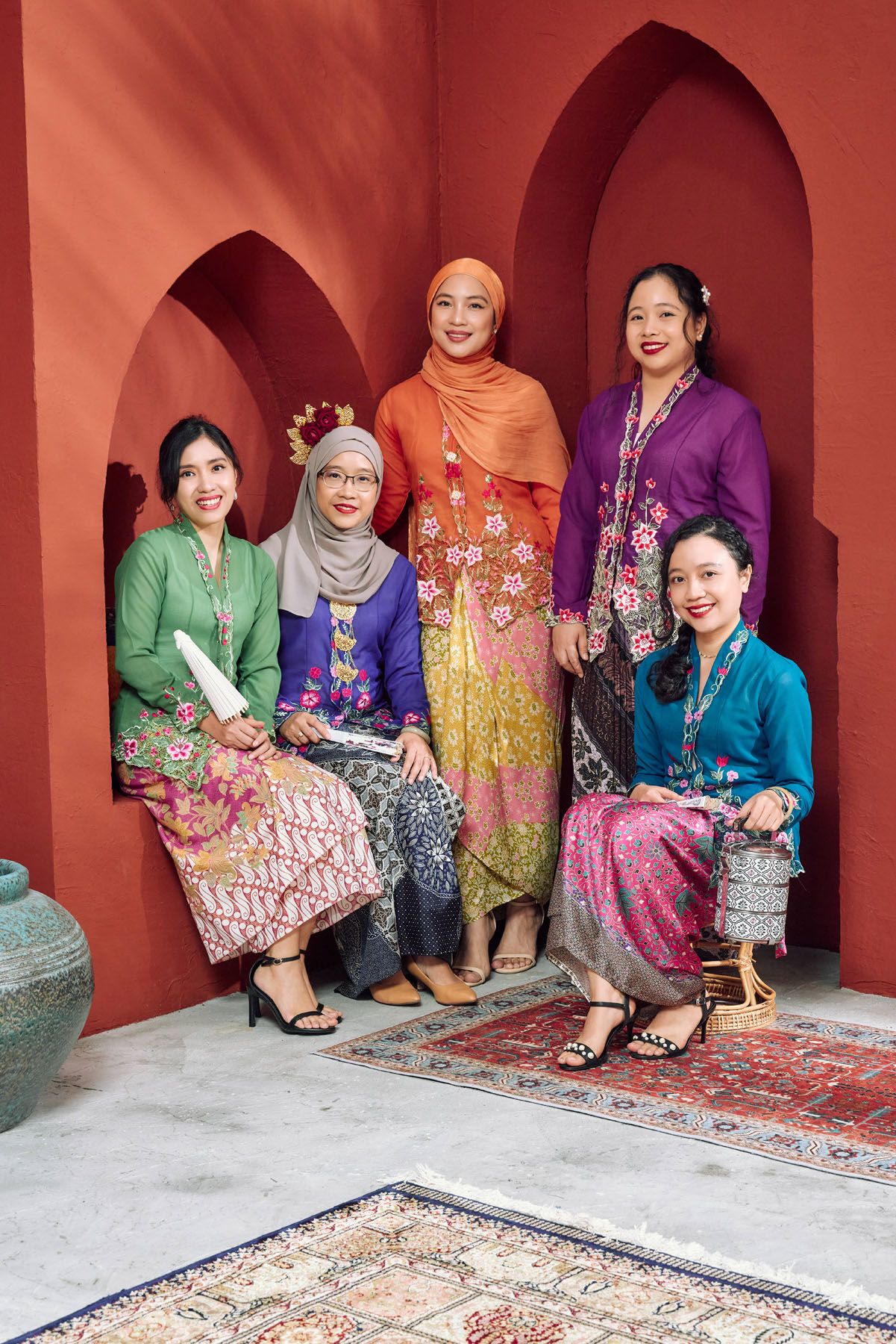 Five women in colorful traditional Malaysian kebaya and batik outfits posing together against a red arched wall.