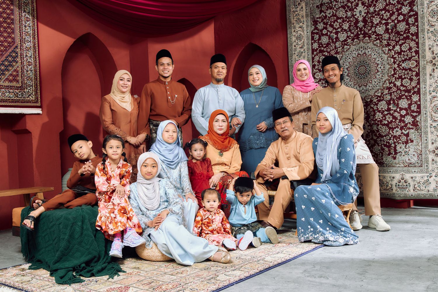 Malaysian family portrait in colorful traditional Raya outfits posed against a red decorative backdrop with ornate carpet.