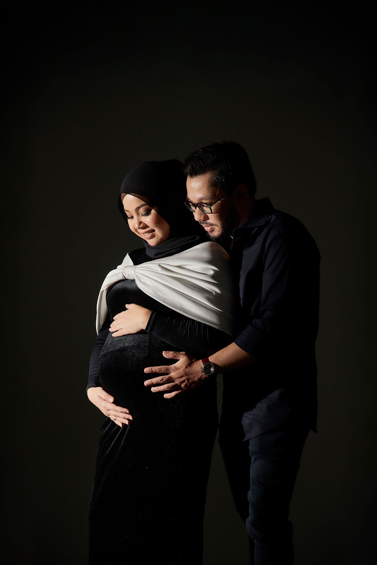 A pregnant Muslim woman in hijab and black dress embraced from behind by her husband, both cradling her baby bump.