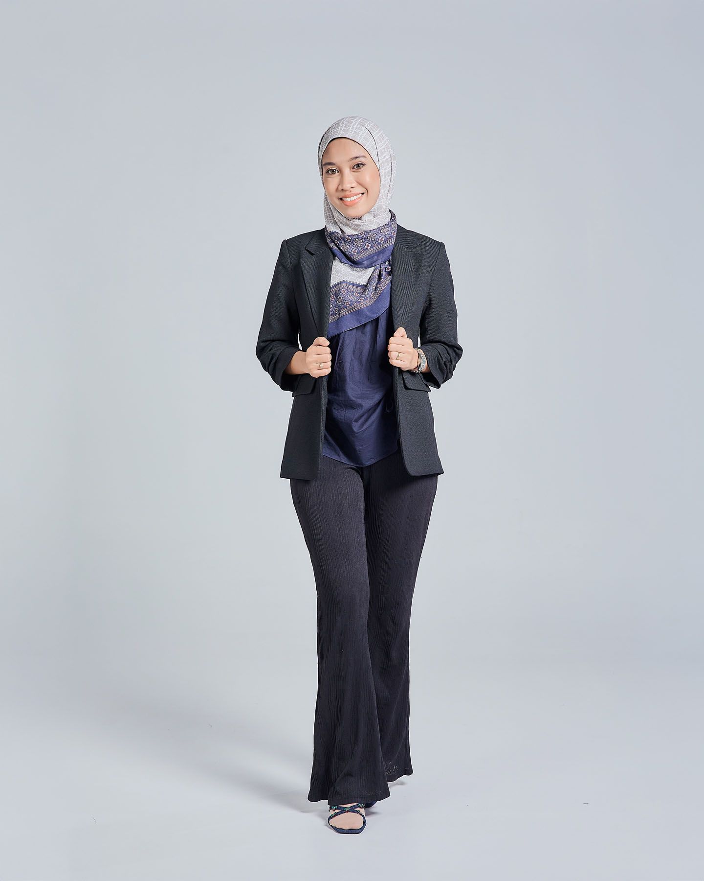 Muslim woman in black blazer, navy top, and wide-leg pants with gray hijab and scarf, giving thumbs up.