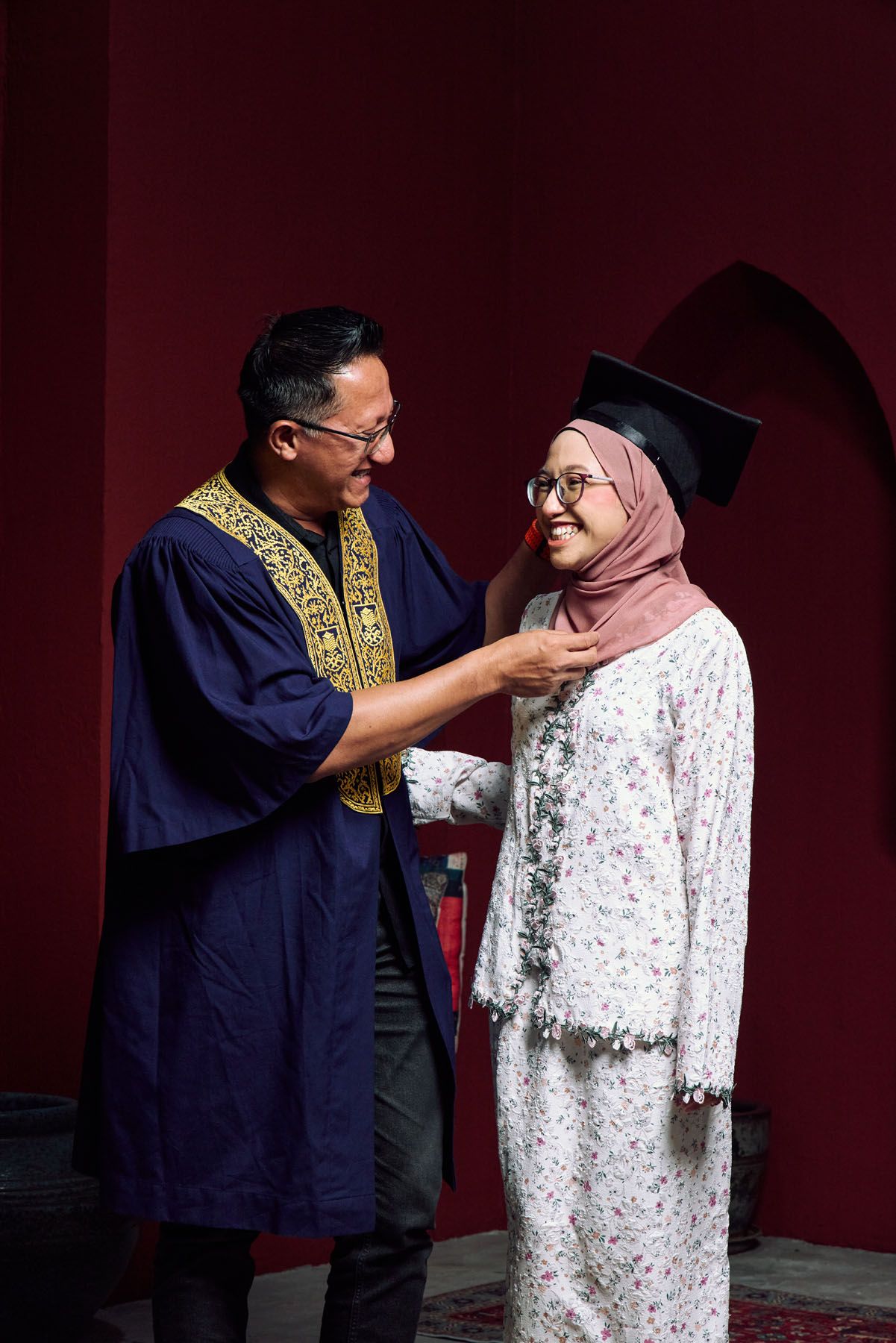 Man in graduation robe adjusts collar of smiling Muslim woman in mortarboard cap.