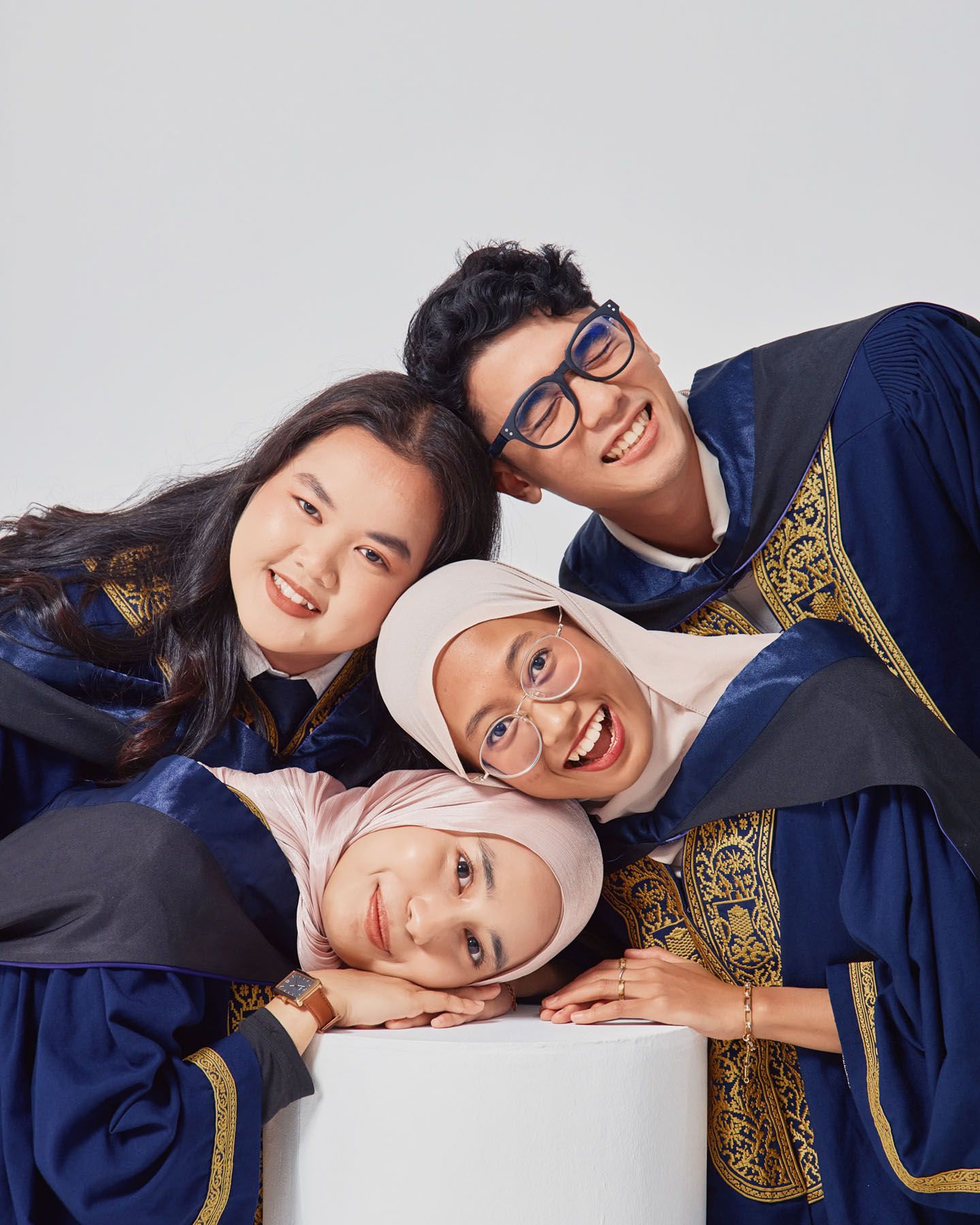 Four smiling graduates in navy robes leaning together over white cylinder for fun group photo.