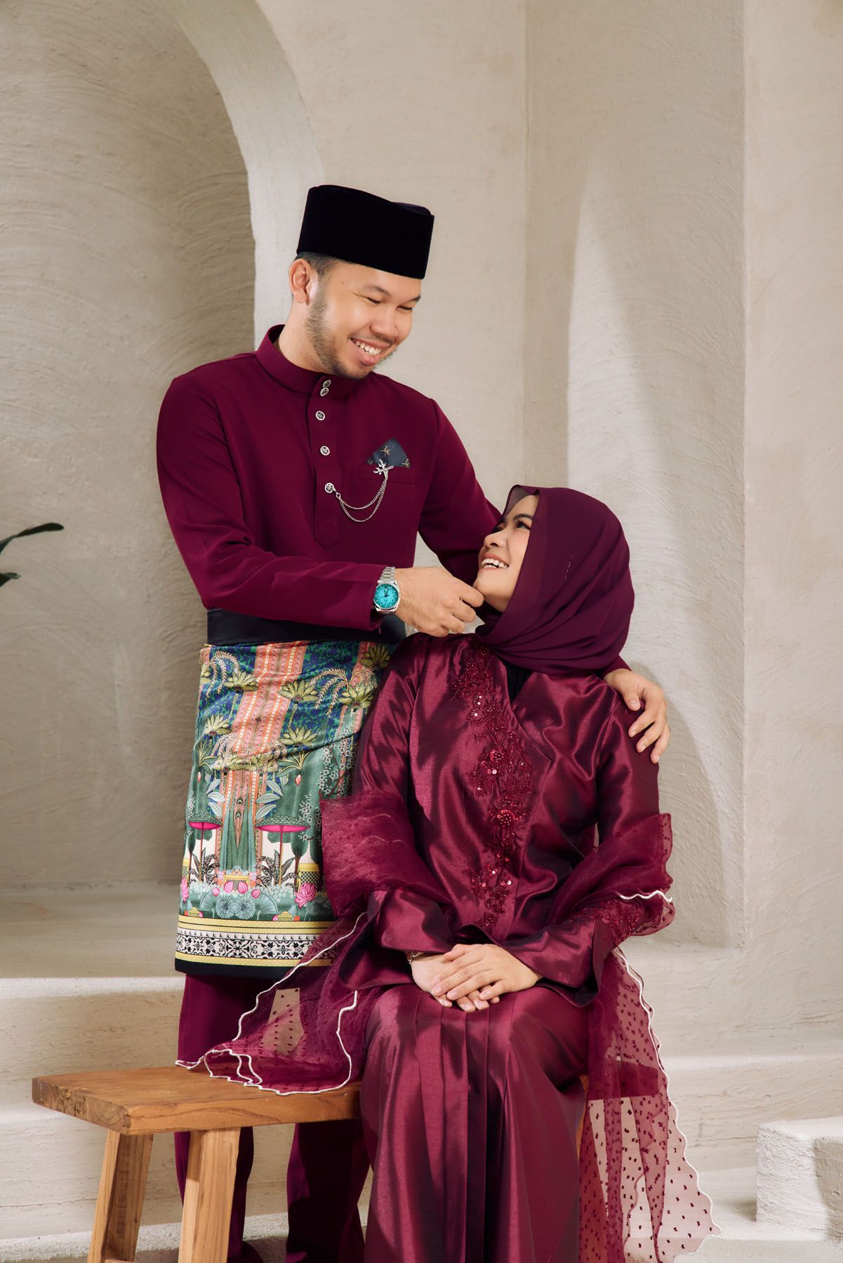 A smiling Malaysian couple in matching maroon Baju Melayu and Baju Kurung traditional outfits posing together indoors.