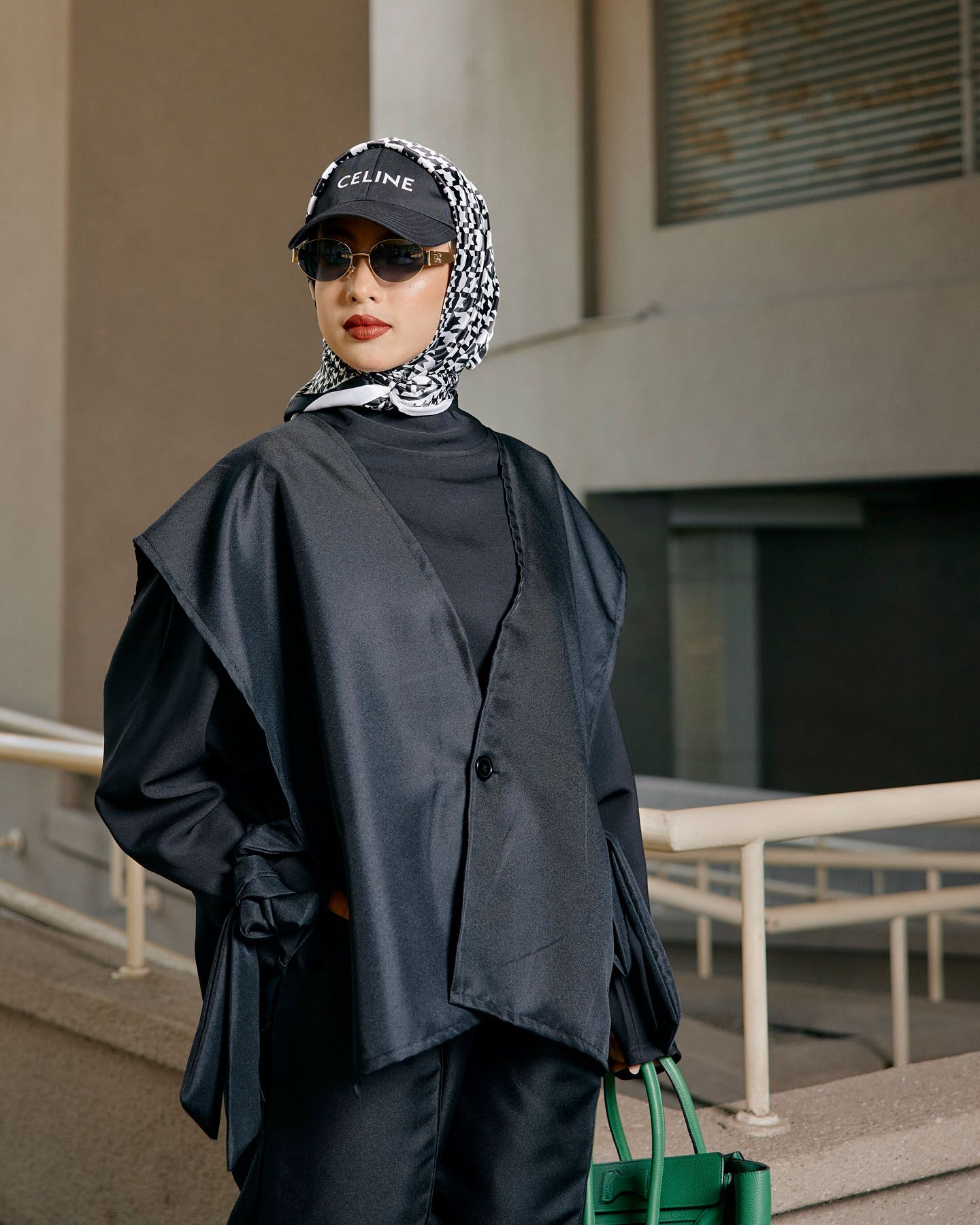 Fashionable woman in black asymmetric jacket and patterned hijab with Celine cap outdoors.