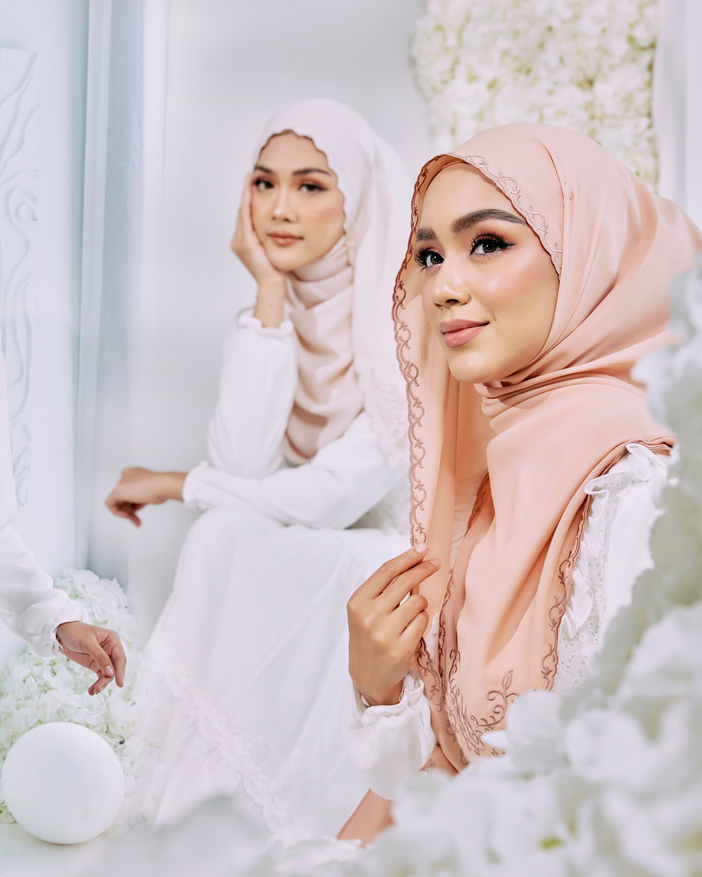 Woman in peach hijab with lace trim posing near a mirror in a white floral studio.
