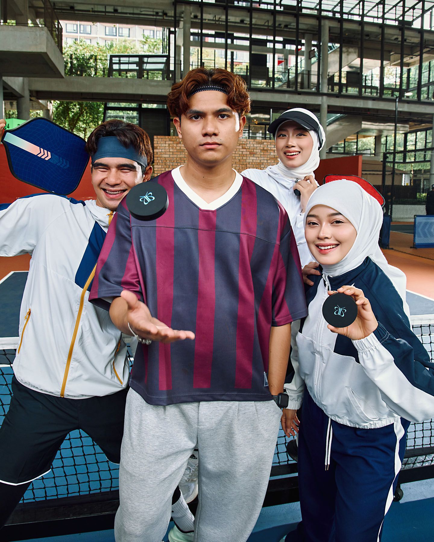 Four young people in sporty outfits holding black compact cosmetics on an outdoor tennis court setting.