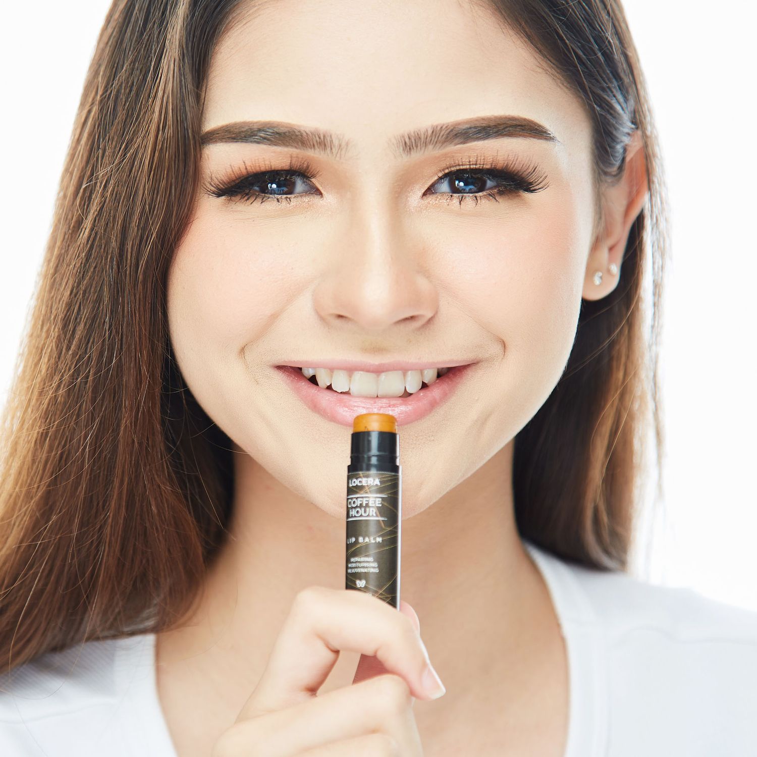 Smiling woman holding a lip balm product close to her lips against a white background.