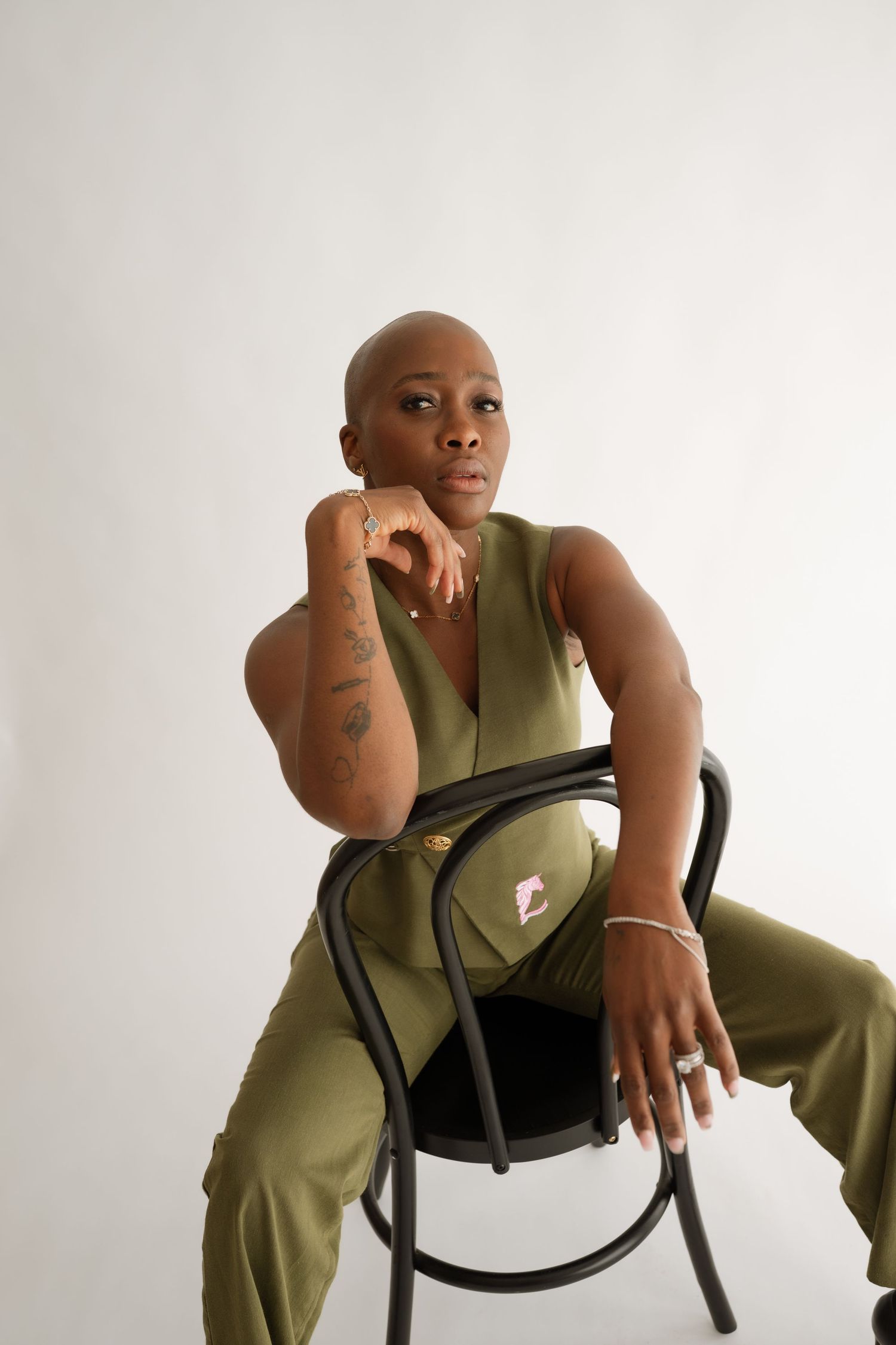 Bald woman in olive green jumpsuit sitting on black chair, showcasing tattoos and jewelry against white background.