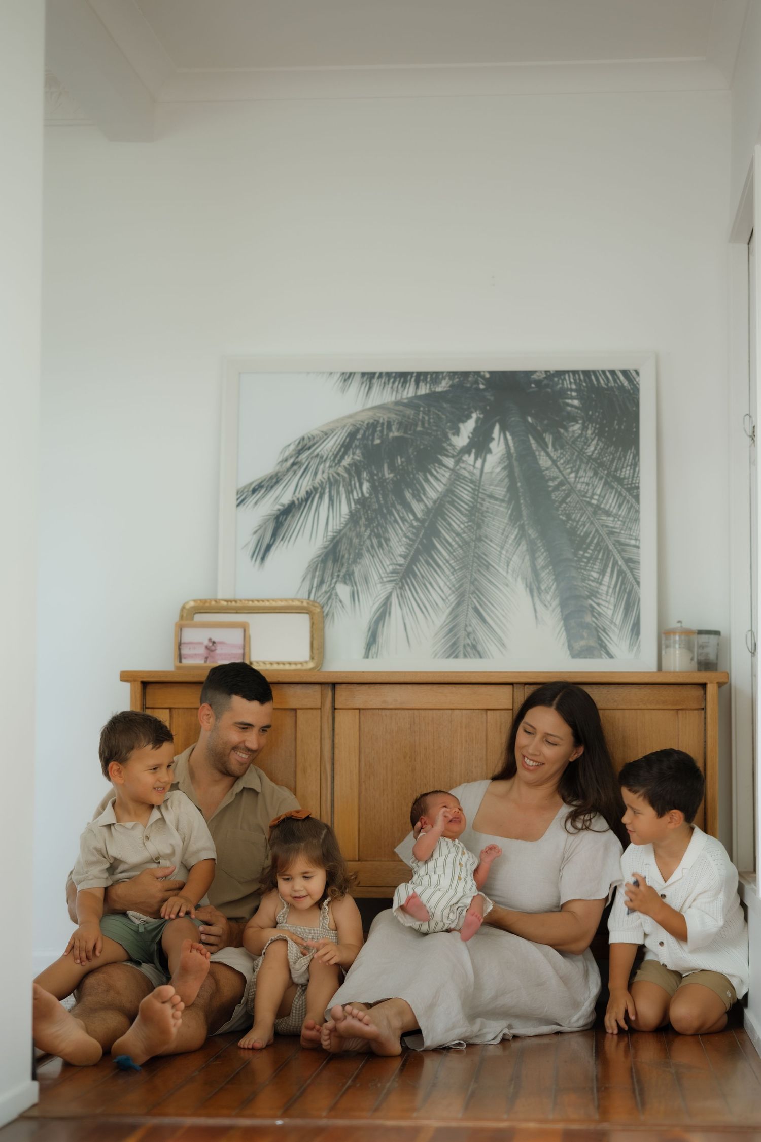 Happy family with newborn baby sitting on floor, parents smiling with three young children near wooden dresser.