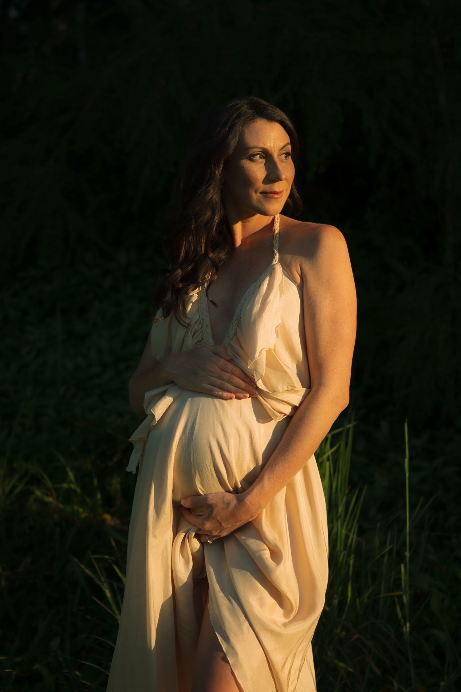 Beautiful pregnant woman in elegant cream halter dress stands in nature during golden hour.