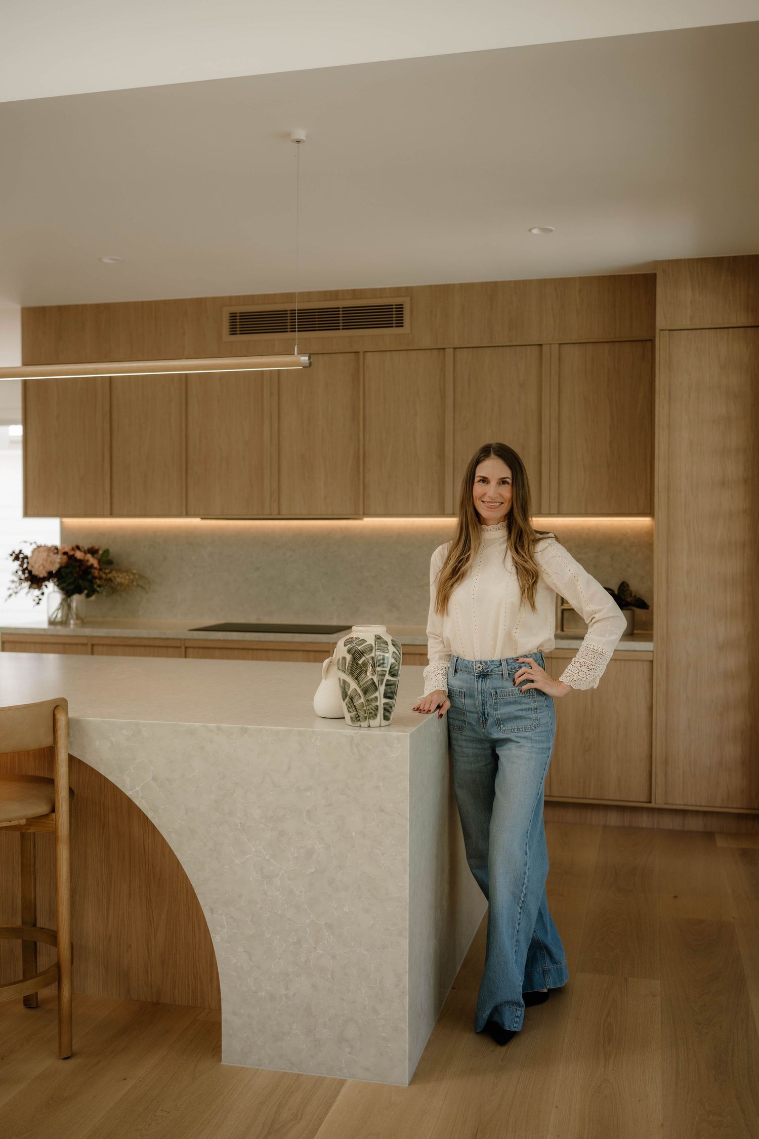 Smiling woman in white top and jeans posing beside a curved kitchen island in modern kitchen.