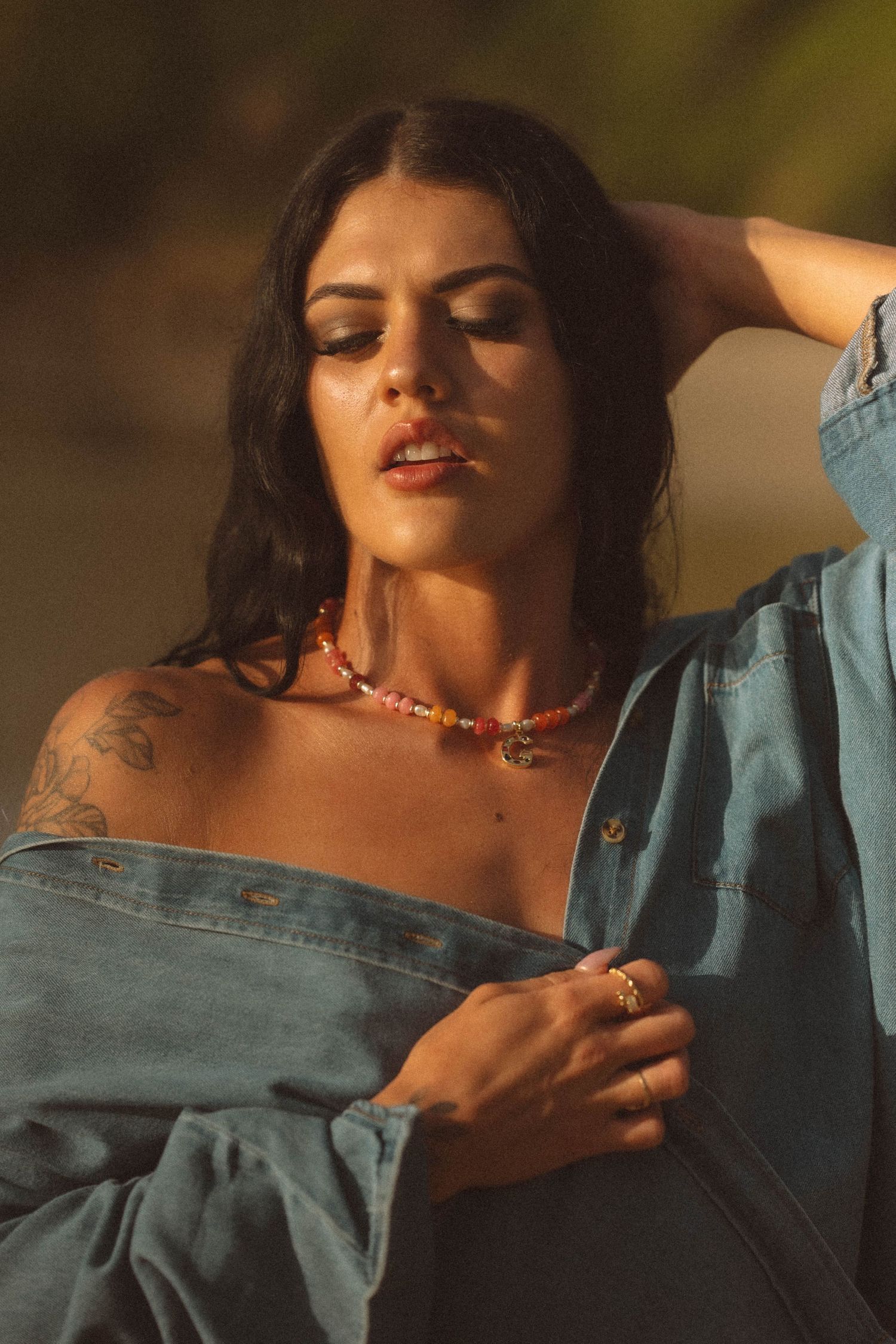Dark-haired woman with tattoos wears colorful beaded necklace in denim shirt at golden hour.