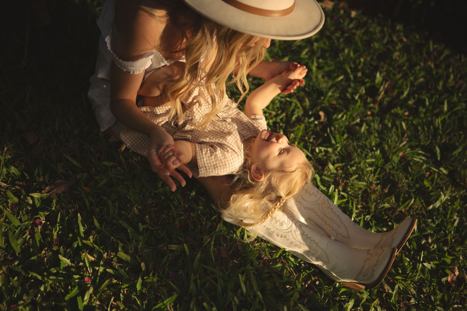 A mother in a straw hat sits on green grass with her blonde toddler daughter in a warm golden light.
