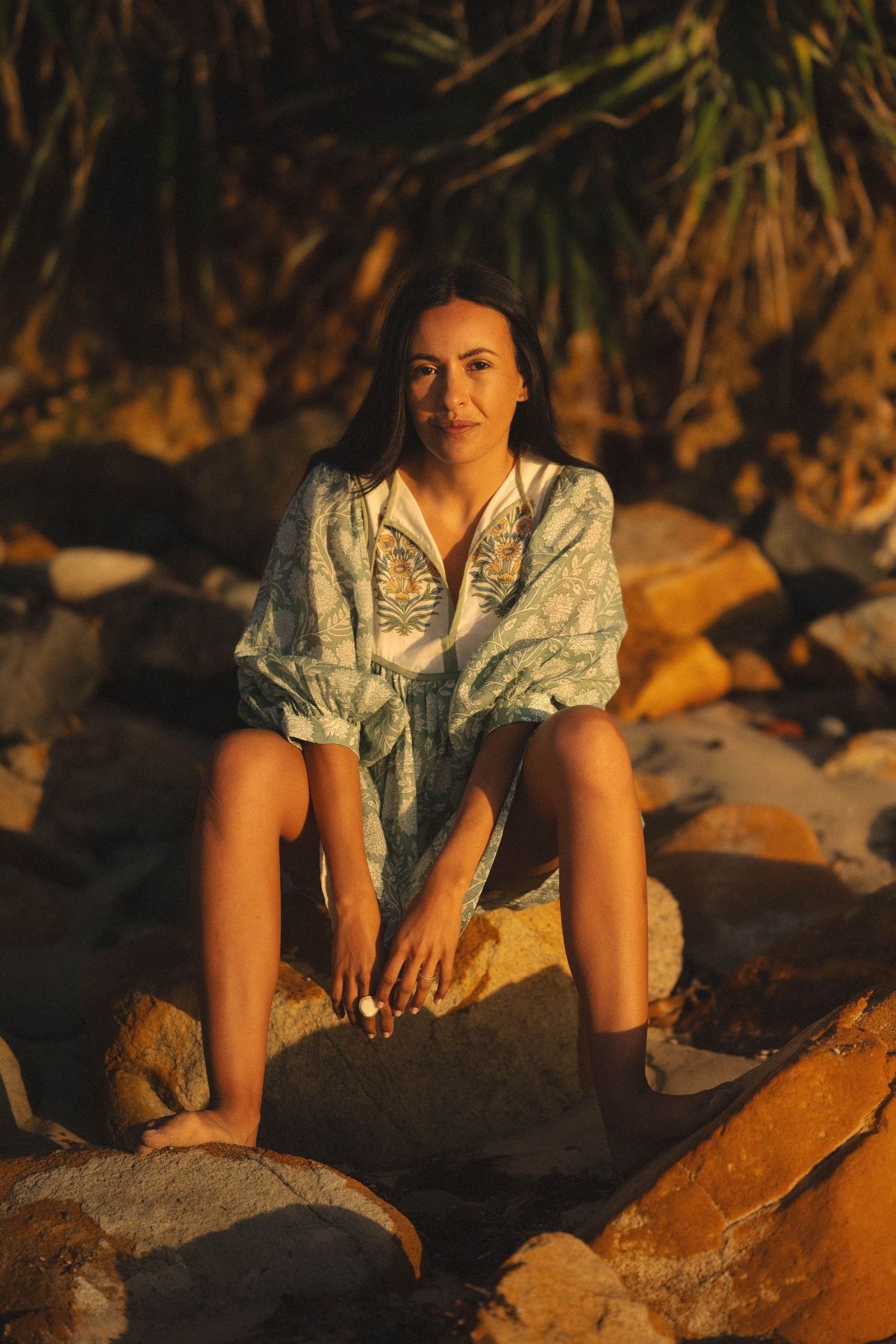 Woman in floral cover-up sitting on rocks at golden hour beach with tropical palms in background.