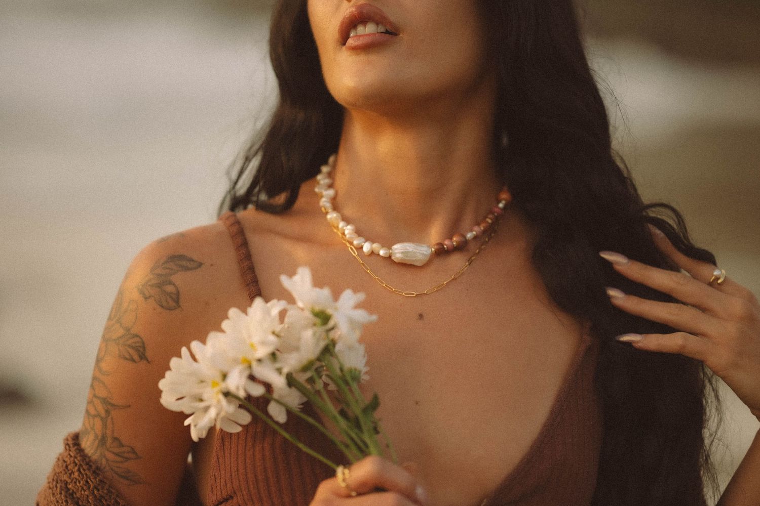 Woman with tattoos wearing pearl necklace holding white flowers at the beach.