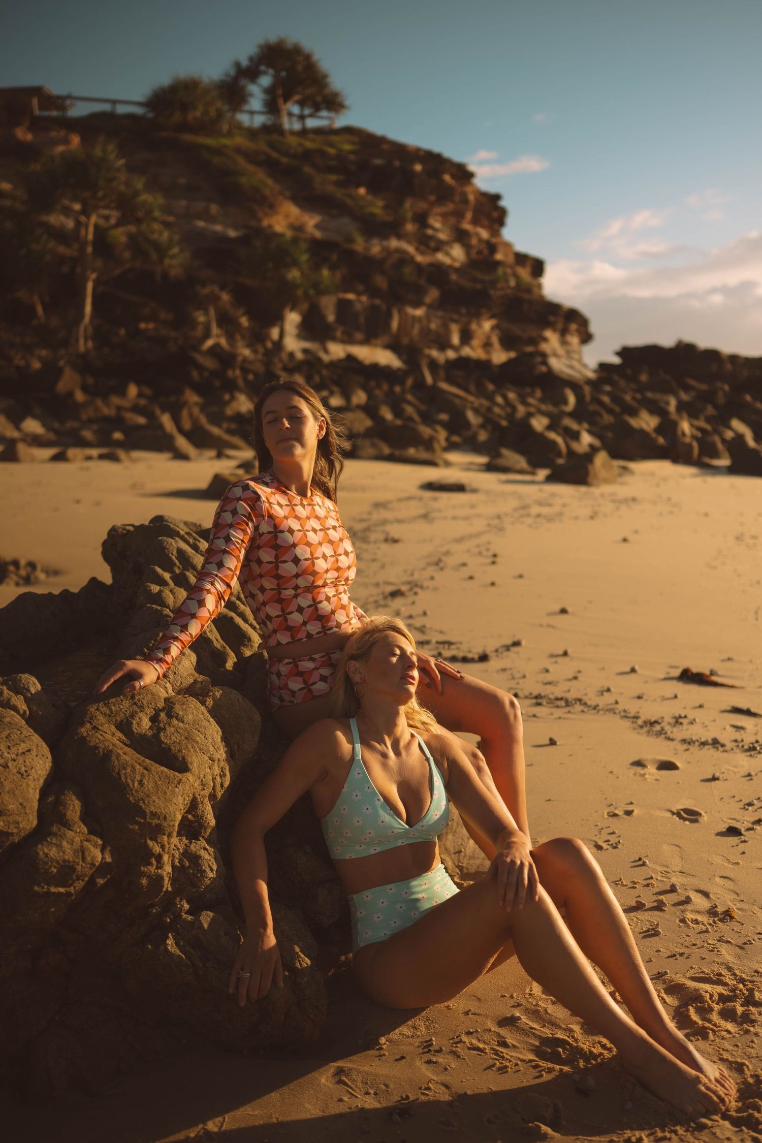Two women in swimwear posing by rocks on sandy beach during golden hour sunset.