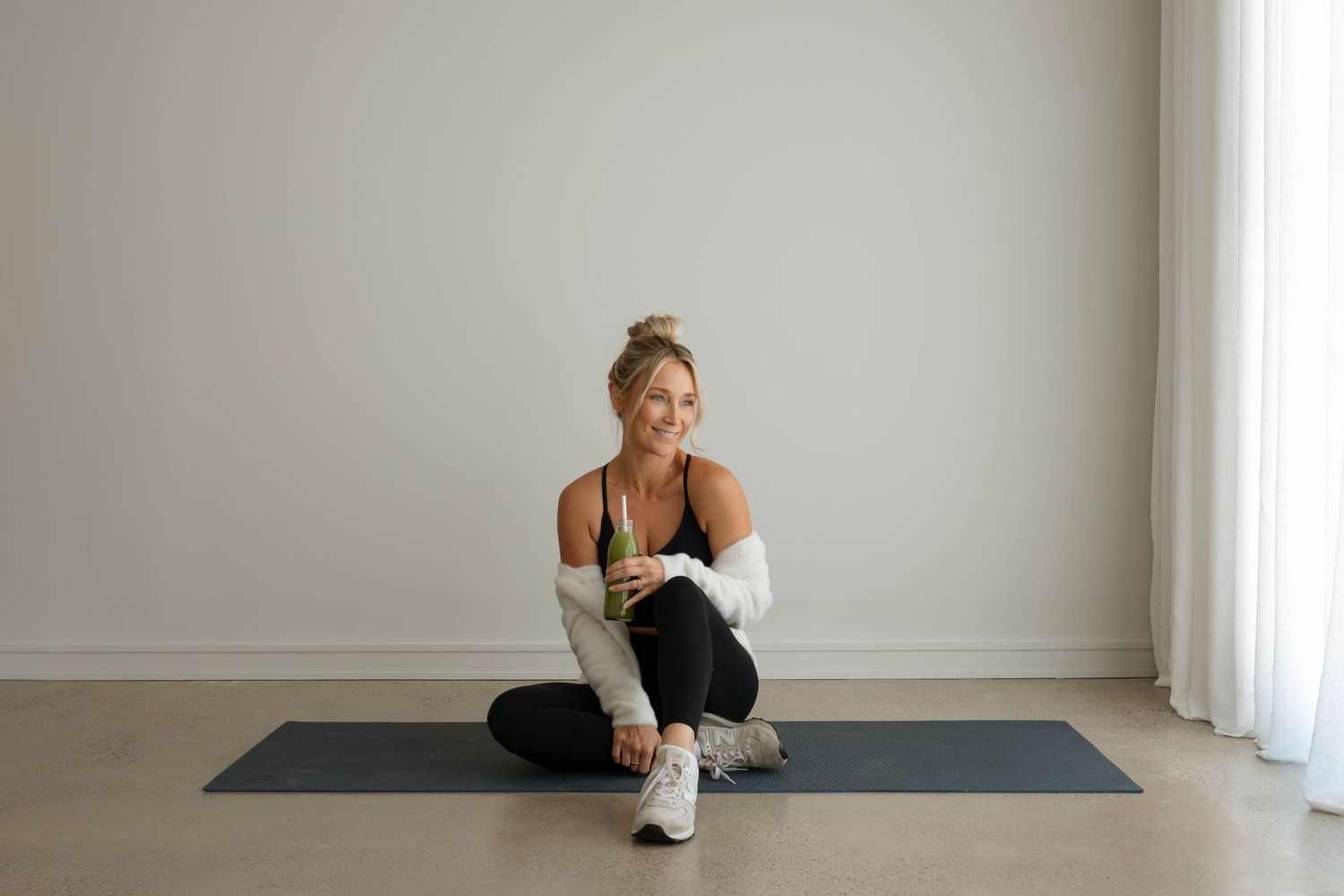 Woman in black sports bra and white cardigan sitting on yoga mat holding green smoothie.
