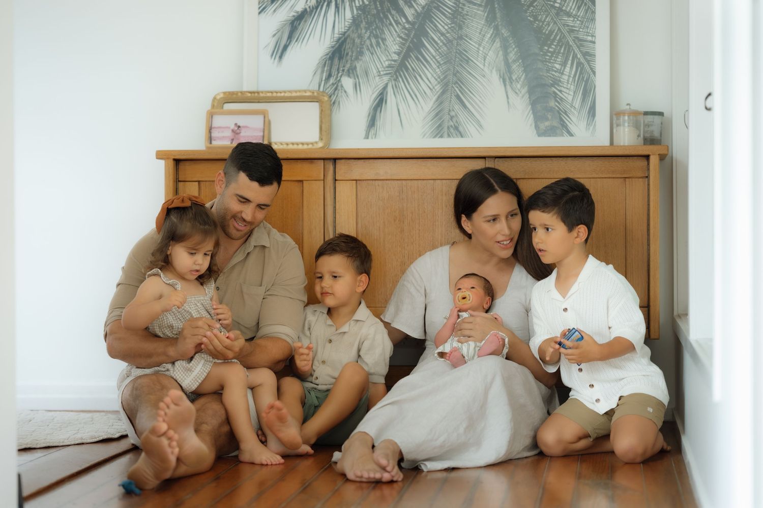Family of six sitting on hardwood floor near wooden dresser, mother holding newborn with three young children.