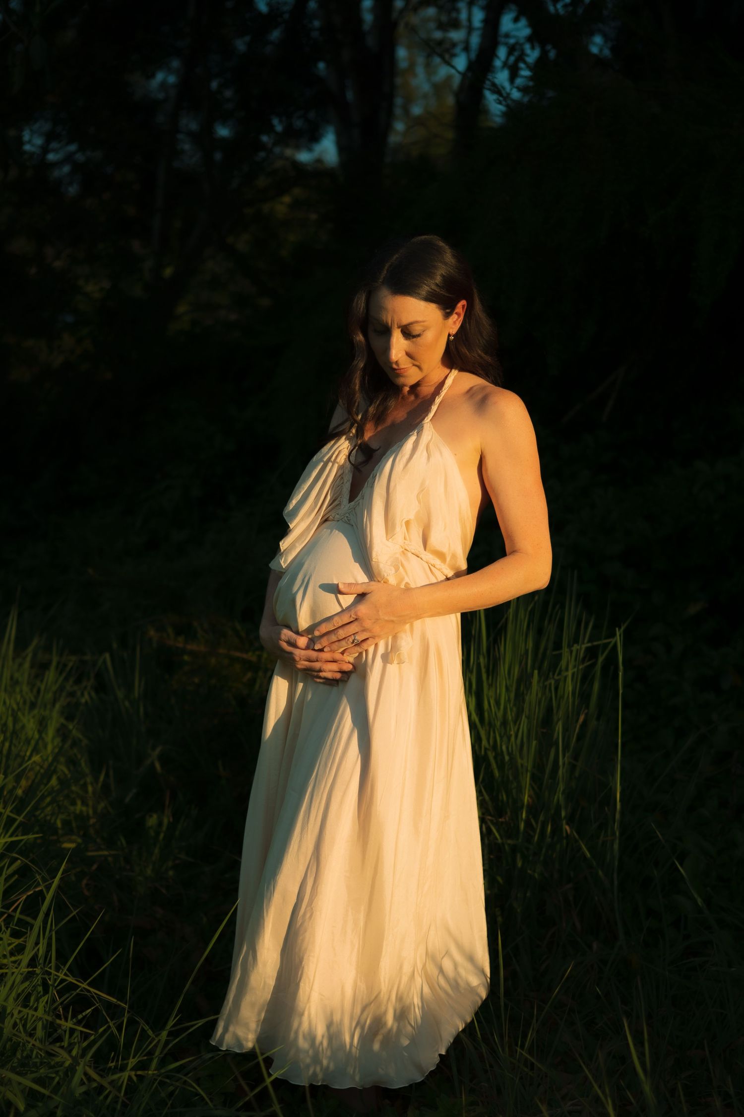 Glowing pregnant woman in white dress gently holds baby bump while standing in sunset meadow.