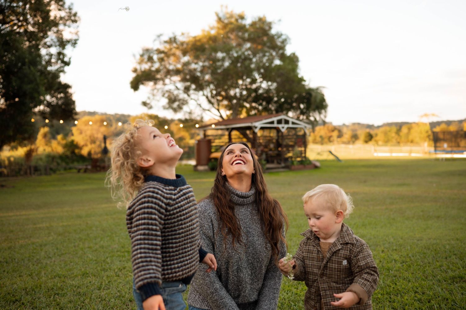 A laughing mother plays outdoors with two young children on a green lawn at golden hour.