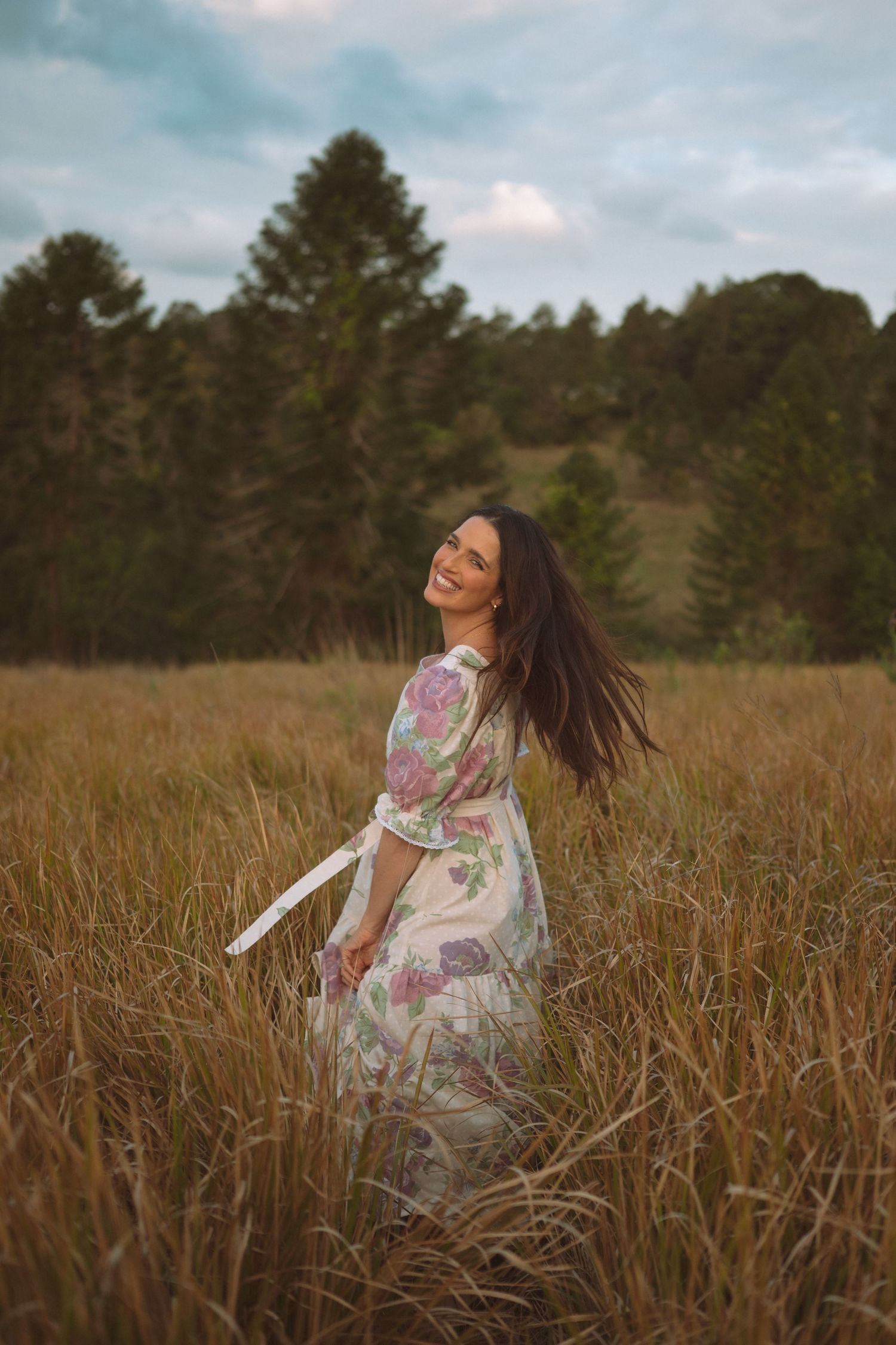 Smiling woman in floral dress twirling in golden meadow with pine trees backdrop.