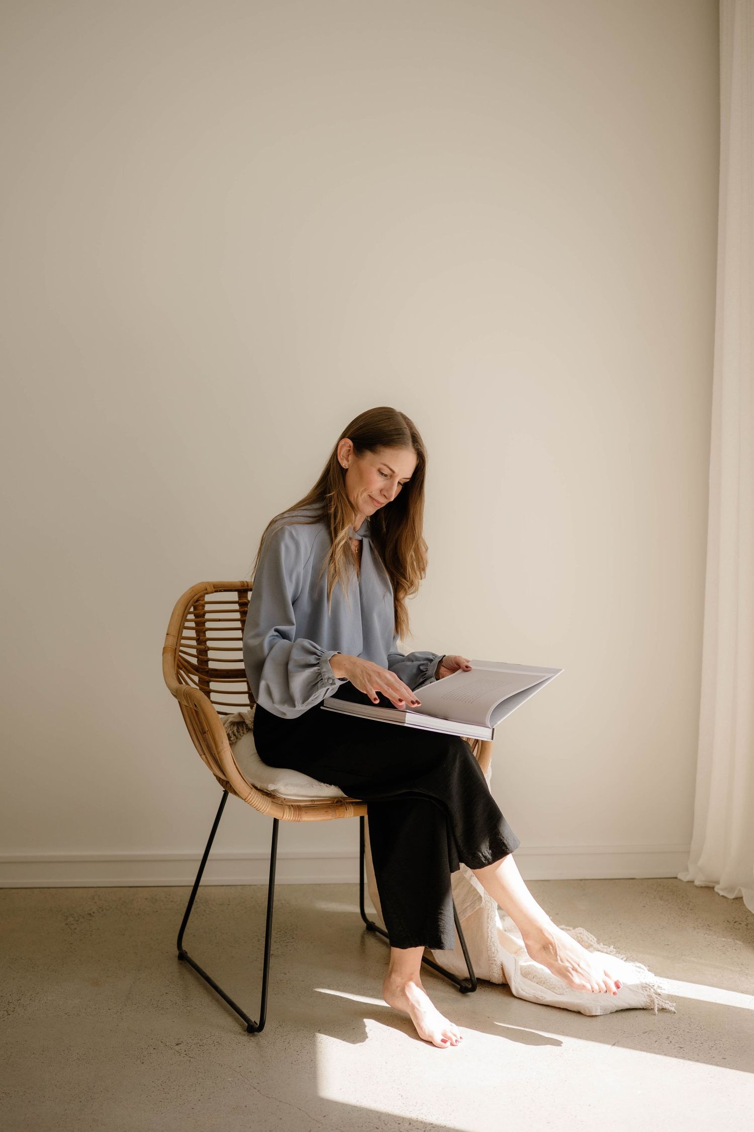 Woman in grey blouse sitting in rattan chair reviewing a large photo album in a sunlit minimal room.