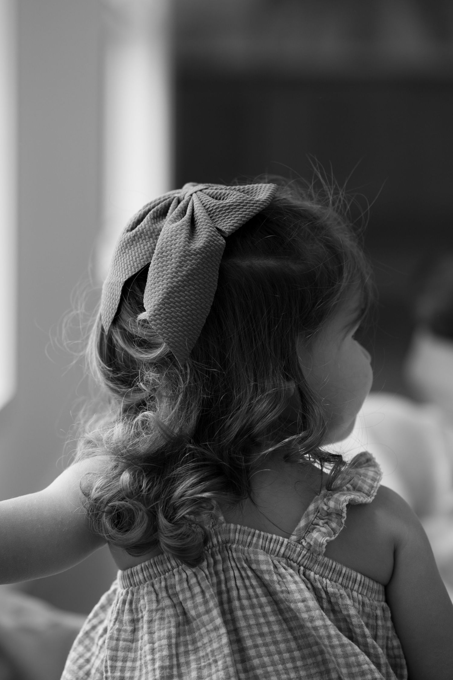 Black and white portrait of curly-haired toddler girl with bow headband in checkered dress.