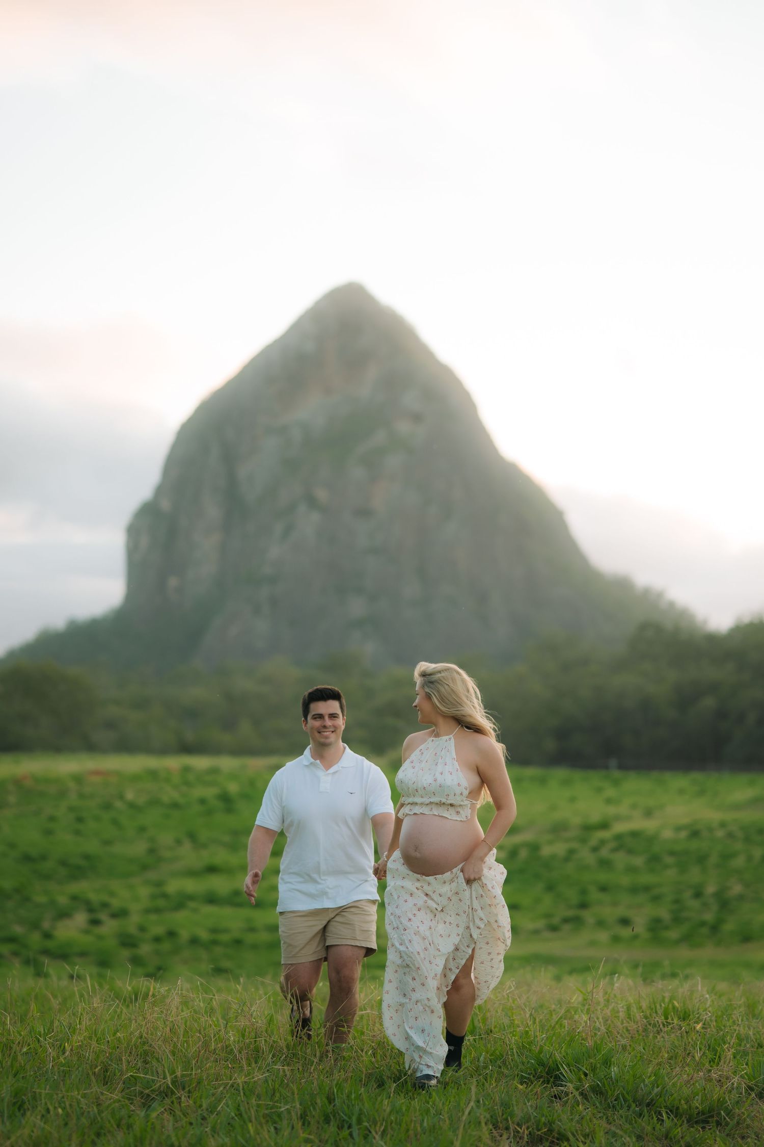 Smiling couple walks hand in hand through green meadow with prominent rocky mountain peak in the background.