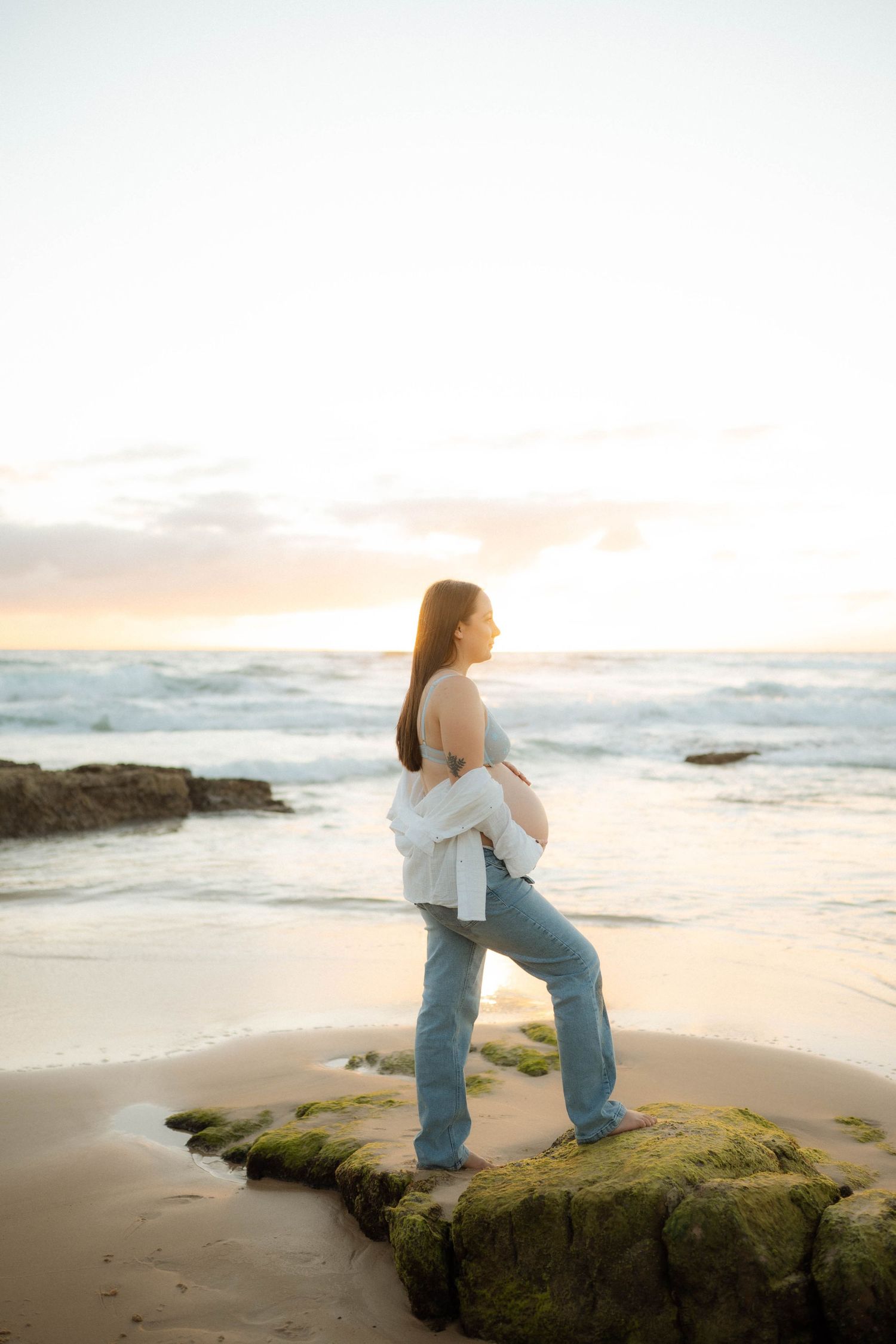 Pregnant woman standing on mossy beach rocks at golden sunset holding her baby bump.