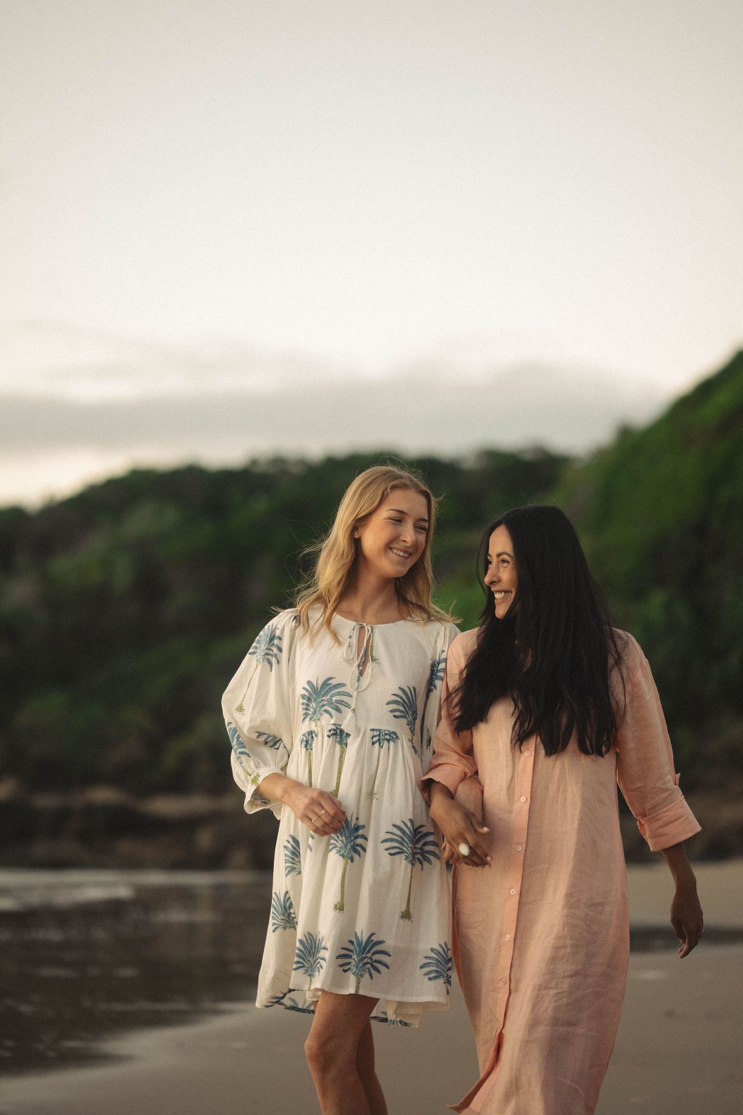 Two women in summer dresses walk together on a beach, smiling and enjoying a coastal sunset.