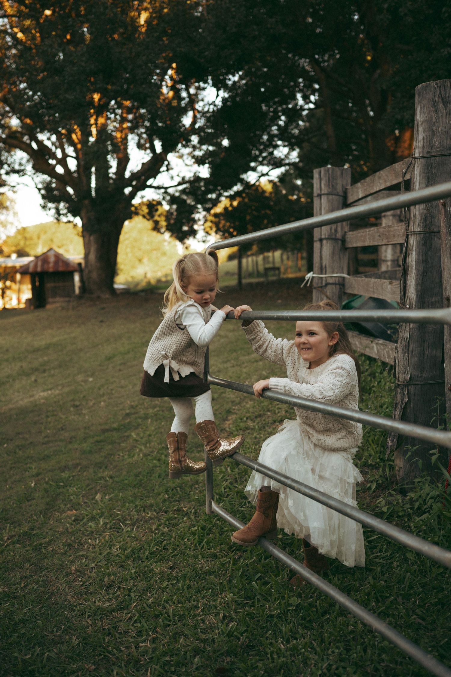 Two young girls in cowboy boots playing on metal farm gate in rustic countryside setting.