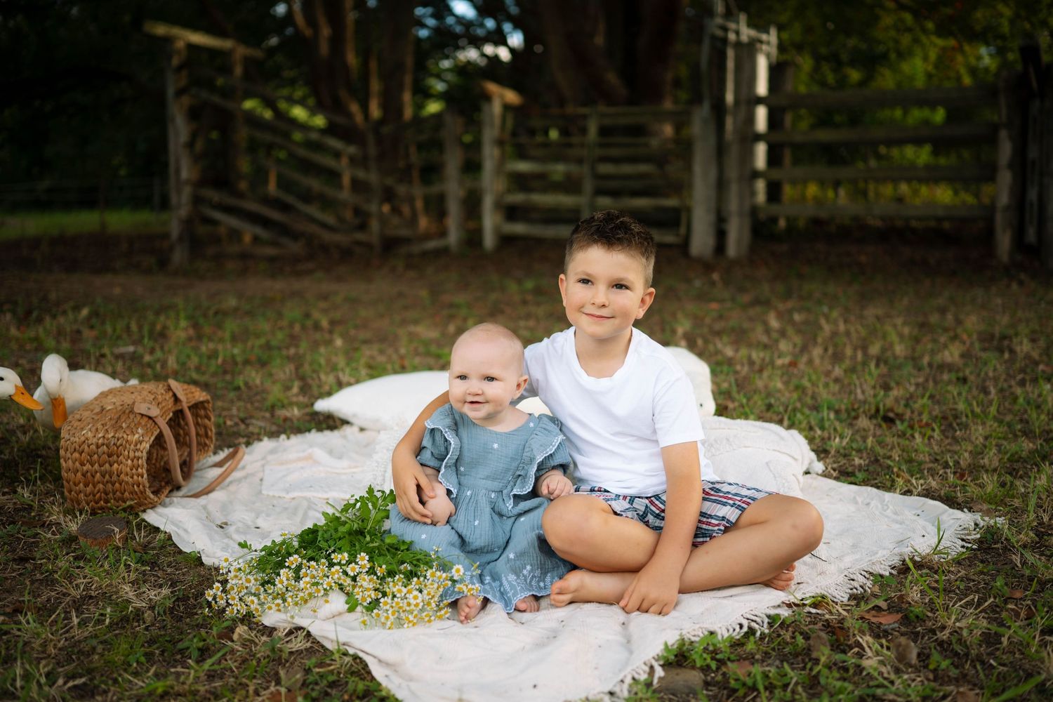 Young boy in white shirt sitting with baby sibling in blue outfit on blanket with wildflowers on a farm.