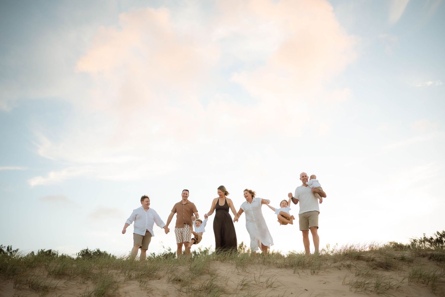 Six family members standing on sand dunes against pastel sunset sky at beach.
